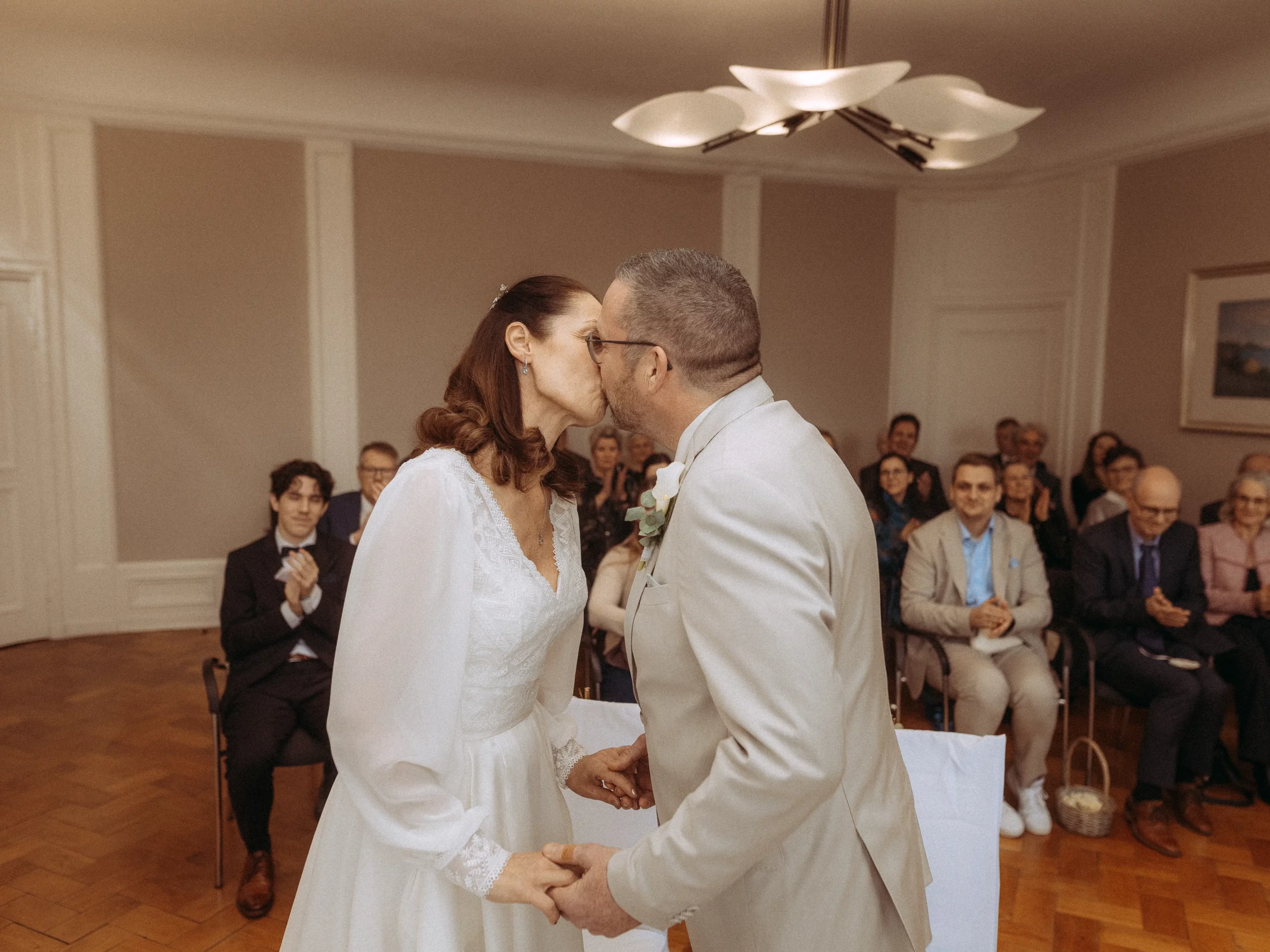 Braut und Bräutigam küssen sich bei ihrer Hochzeitszeremonie, während die Gäste im Saal zusehen, klatschen und lächeln. Wunderschön eingefangen von Hochzeitsfotograf MV.