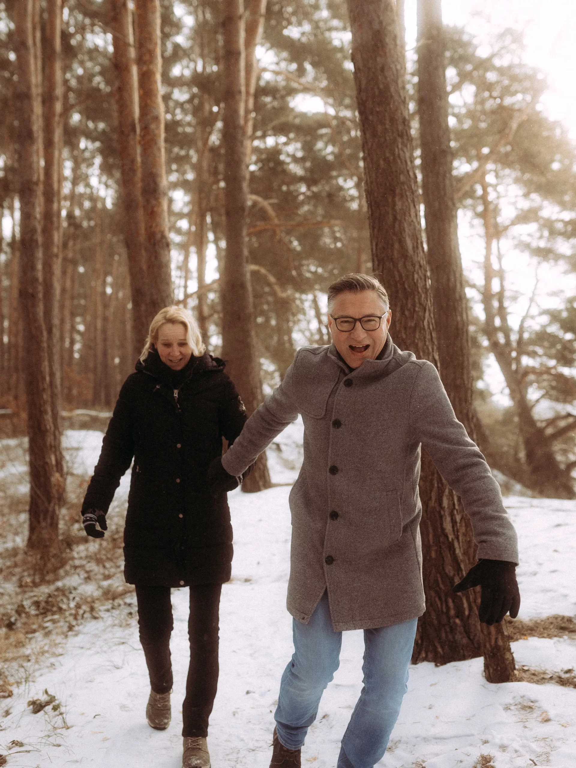Ein Mann und eine Frau gehen Hand in Hand durch einen verschneiten Wald. Der Mann in einem grauen Mantel und mit Brille lacht, während die Frau in einem schwarzen Mantel hinter ihm lächelt. Hohe Bäume umgeben das Paar - perfekt für Hochzeitsfotos MV in dieser zauberhaften Winterlandschaft.