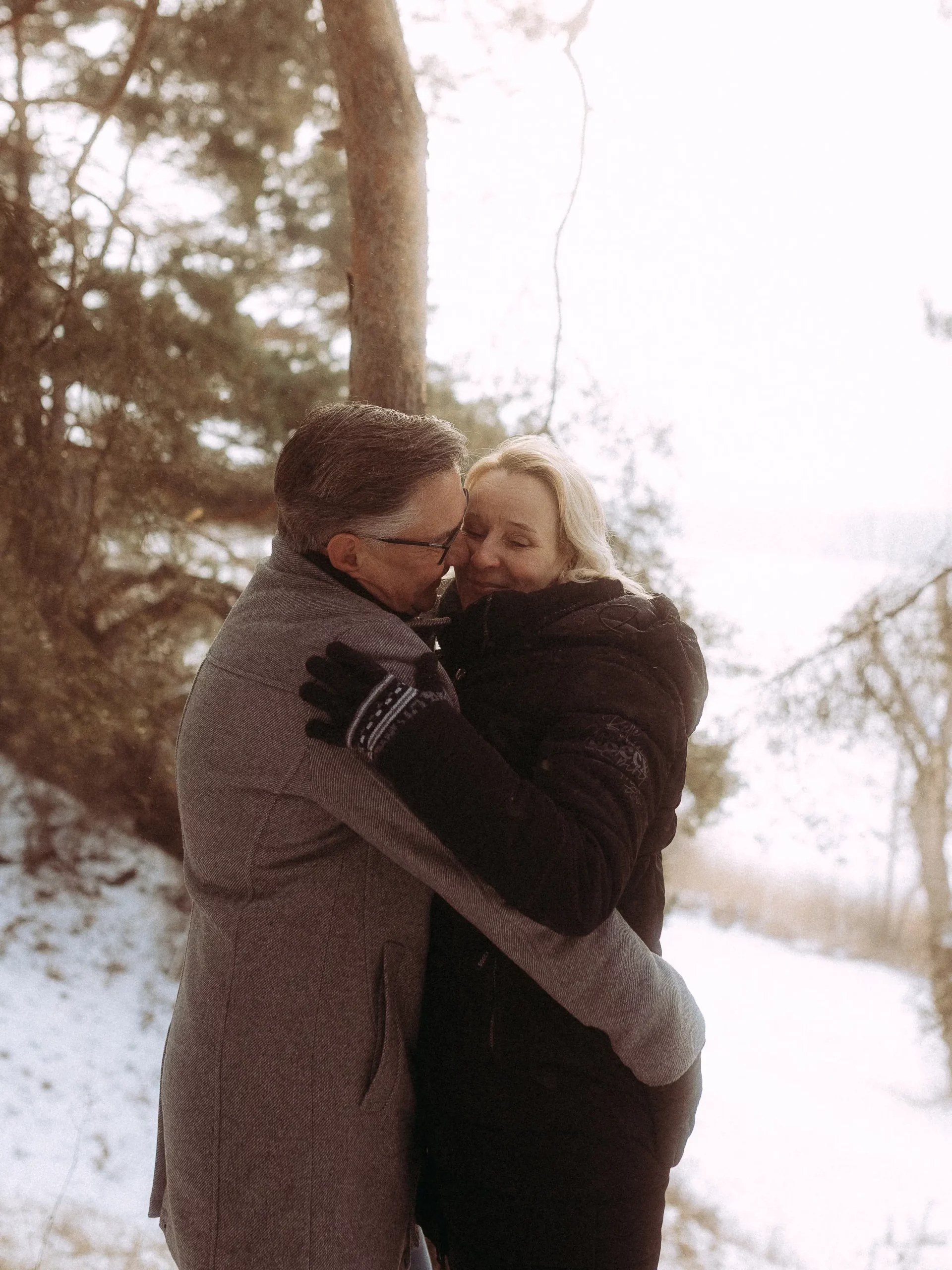 Ein Paar in Wintermantel und Handschuhen umarmt sich im Freien, umgeben von Schnee und Bäumen, mit sanftem Sonnenlicht im Hintergrund - aufgenommen von Fotograf MV für zeitlose Hochzeitsfotos MV.