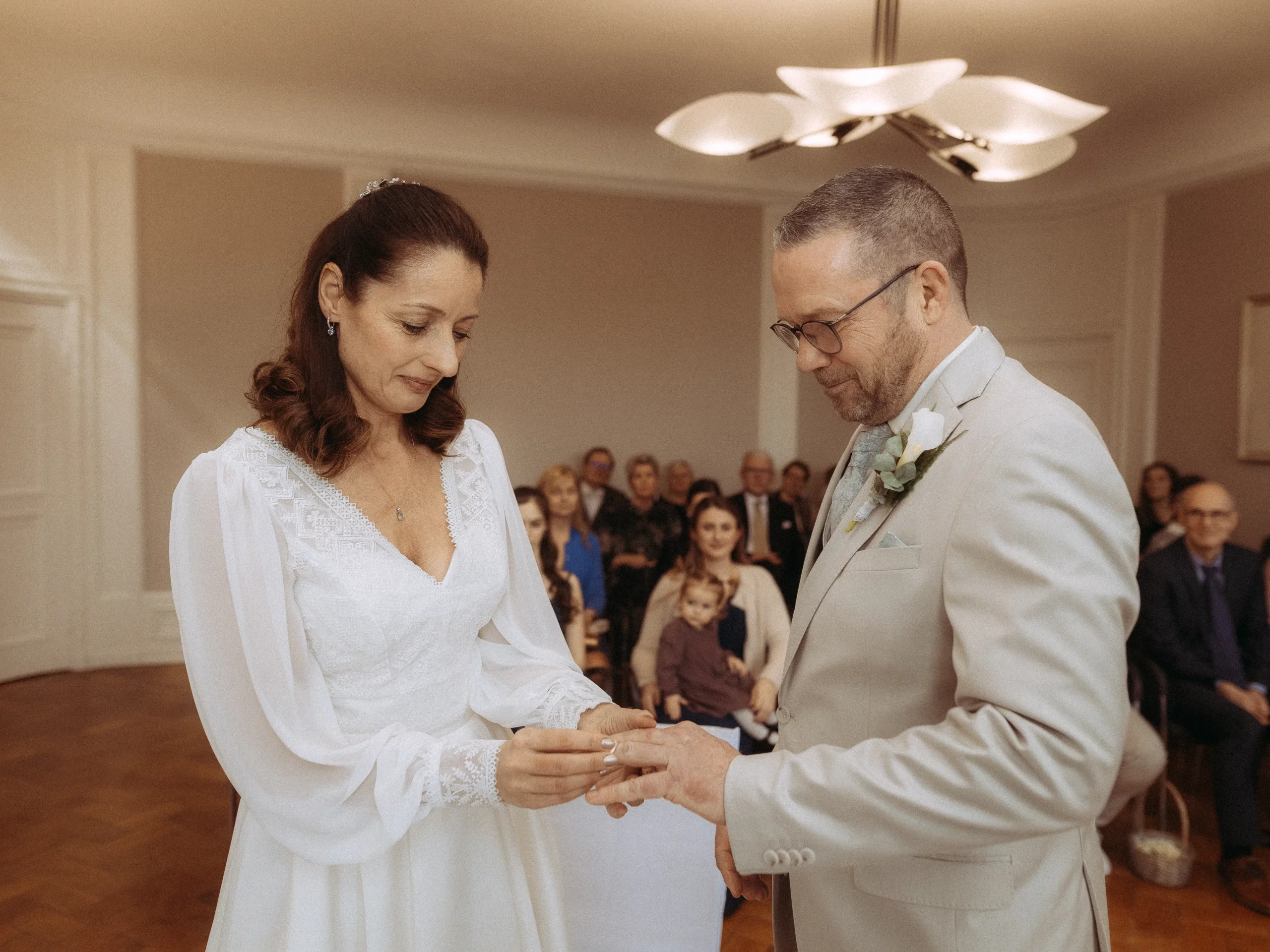Eine Braut in einem weißen Kleid steckt dem Bräutigam während einer Hochzeitszeremonie einen Ring an den Finger, während die Gäste in einem sanft beleuchteten Raum zusehen - wunderschön eingefangen von Hochzeitsfotos MV.