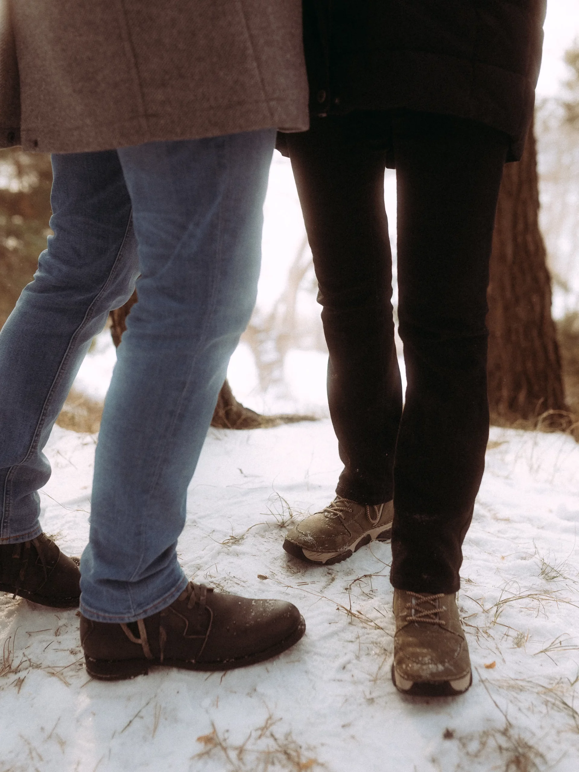 Zwei Personen stehen dicht beieinander im Freien auf schneebedecktem Boden; eine in blauen Jeans und dunklen Schuhen, die andere in schwarzen Hosen und braunen Schuhen. Ihre Oberkörper sind meist außerhalb des Bildausschnitts. Bäume säumen den Hintergrund - aufgenommen von Hochzeitsfotograf MV.