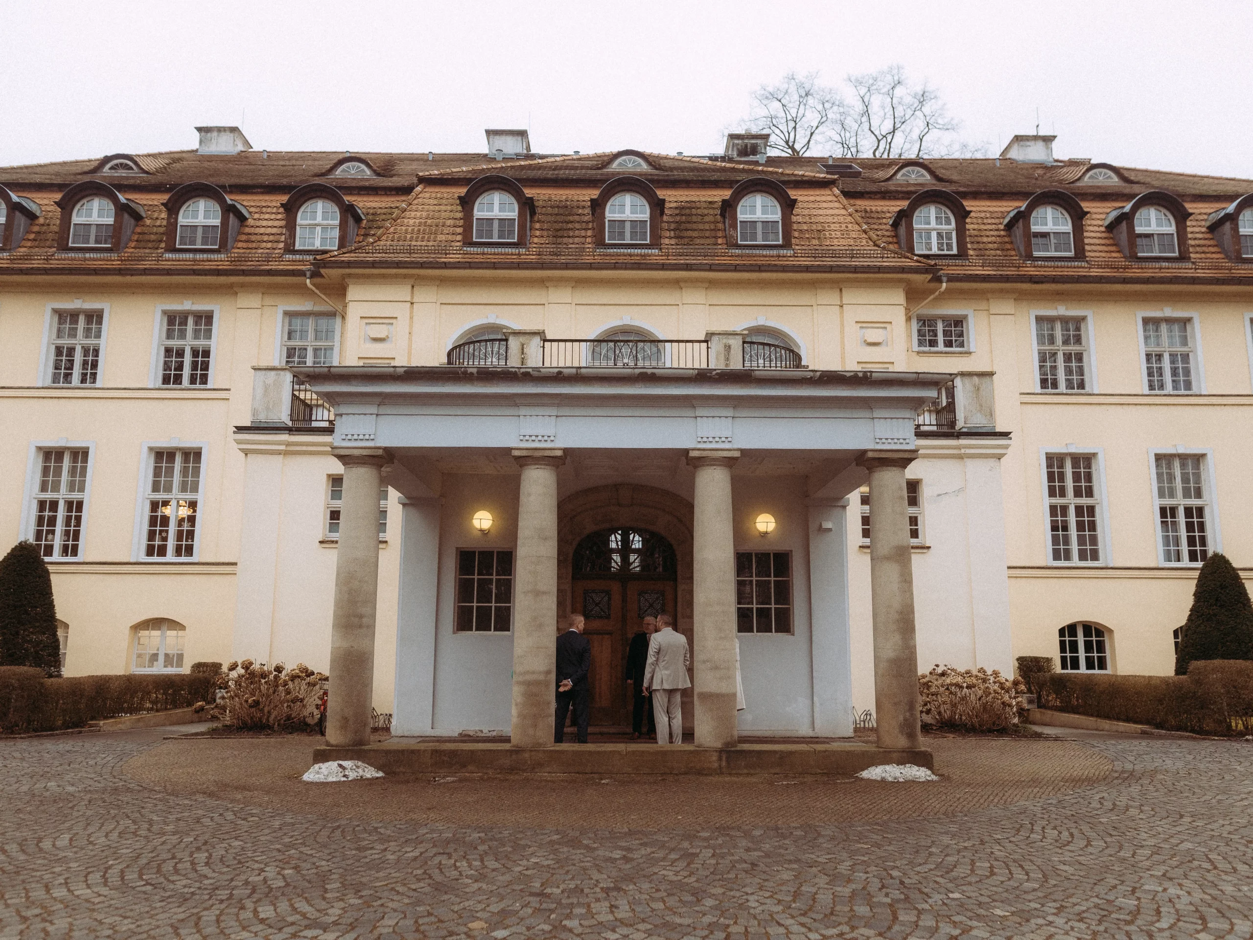 Ein großes, elegantes Gebäude mit einer hellgelben Fassade, Bogenfenstern und einem Ziegeldach. Drei Personen stehen in der Nähe des Eingangs unter einem von Säulen getragenen Laubengang - ideal für einen Fotografen MV, um besondere Momente festzuhalten. Kopfsteinpflaster und Sträucher säumen die Seiten.