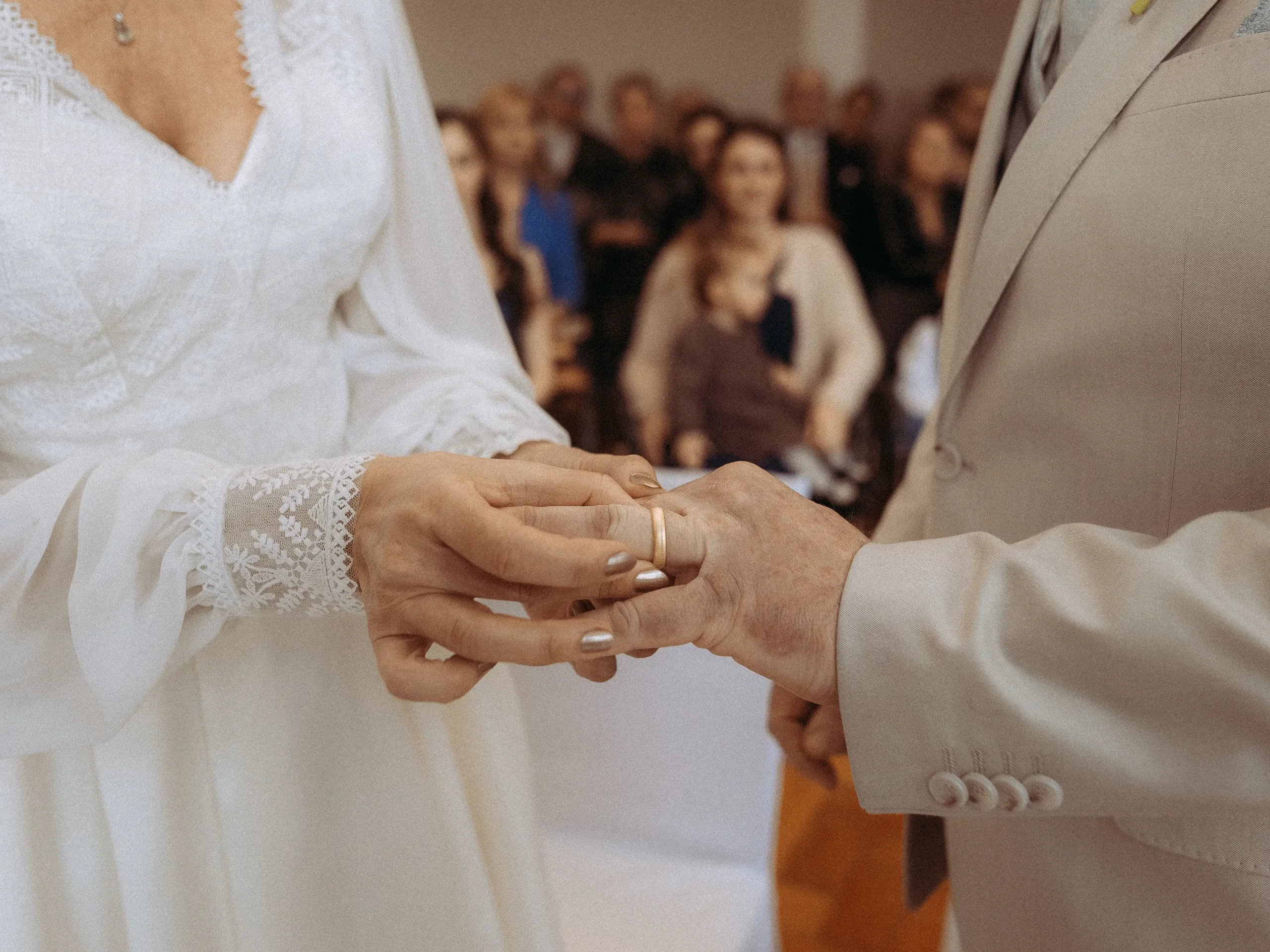 Eine Braut in einem weißen Spitzenkleid steckt dem Bräutigam während der Zeremonie einen Ehering an den Finger, während die Gäste im unscharfen Hintergrund zusehen - wunderschön eingefangen von Hochzeitsfotograf MV.