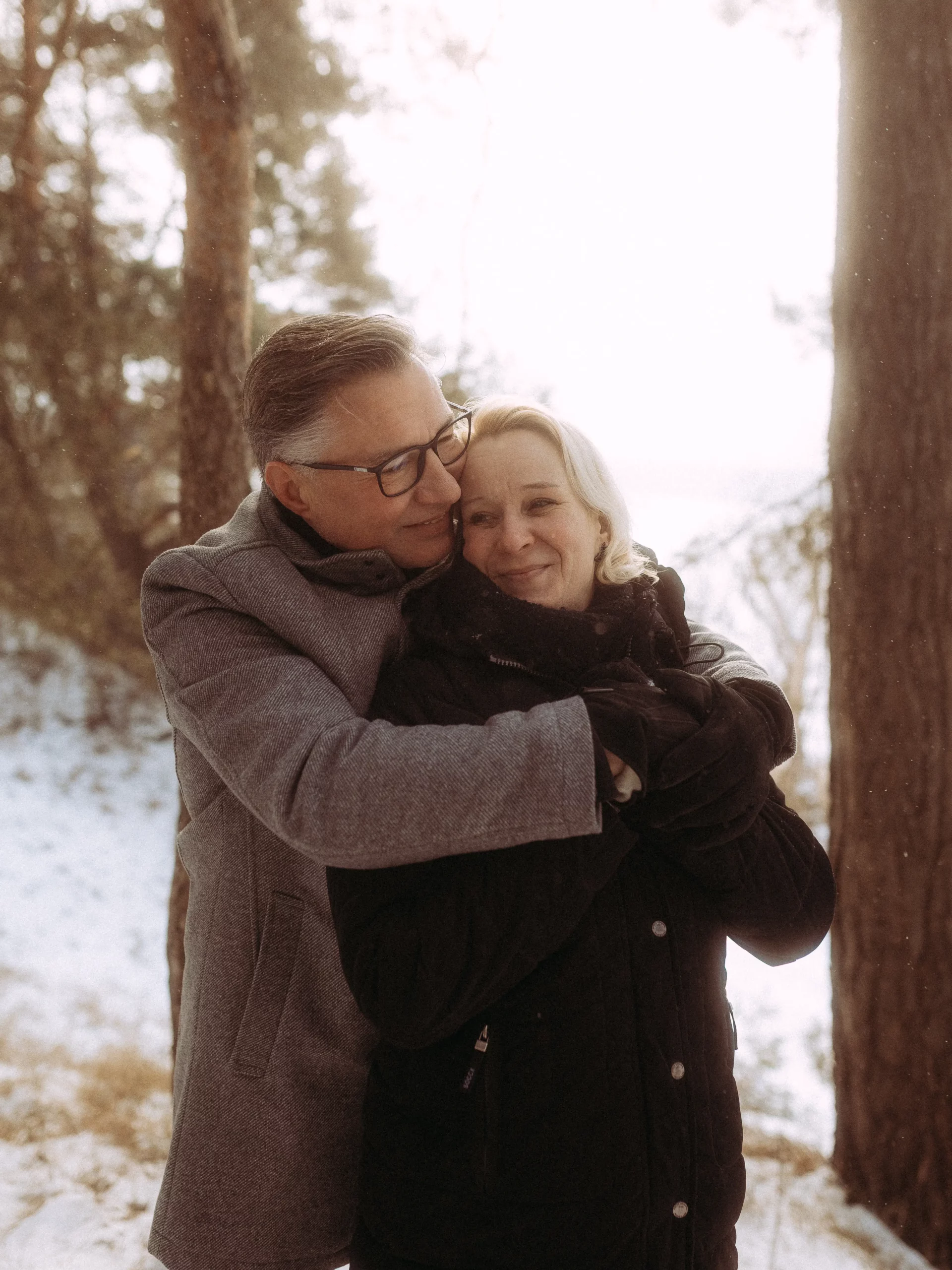 Ein älteres Paar in Wintermänteln steht draußen in einem verschneiten Wald. Der Mann umarmt die Frau von hinten, und beide lächeln warm, während das weiche Sonnenlicht durch die Bäume fällt. Aufgenommen von Fotograf MV für zeitlose Erinnerungen.