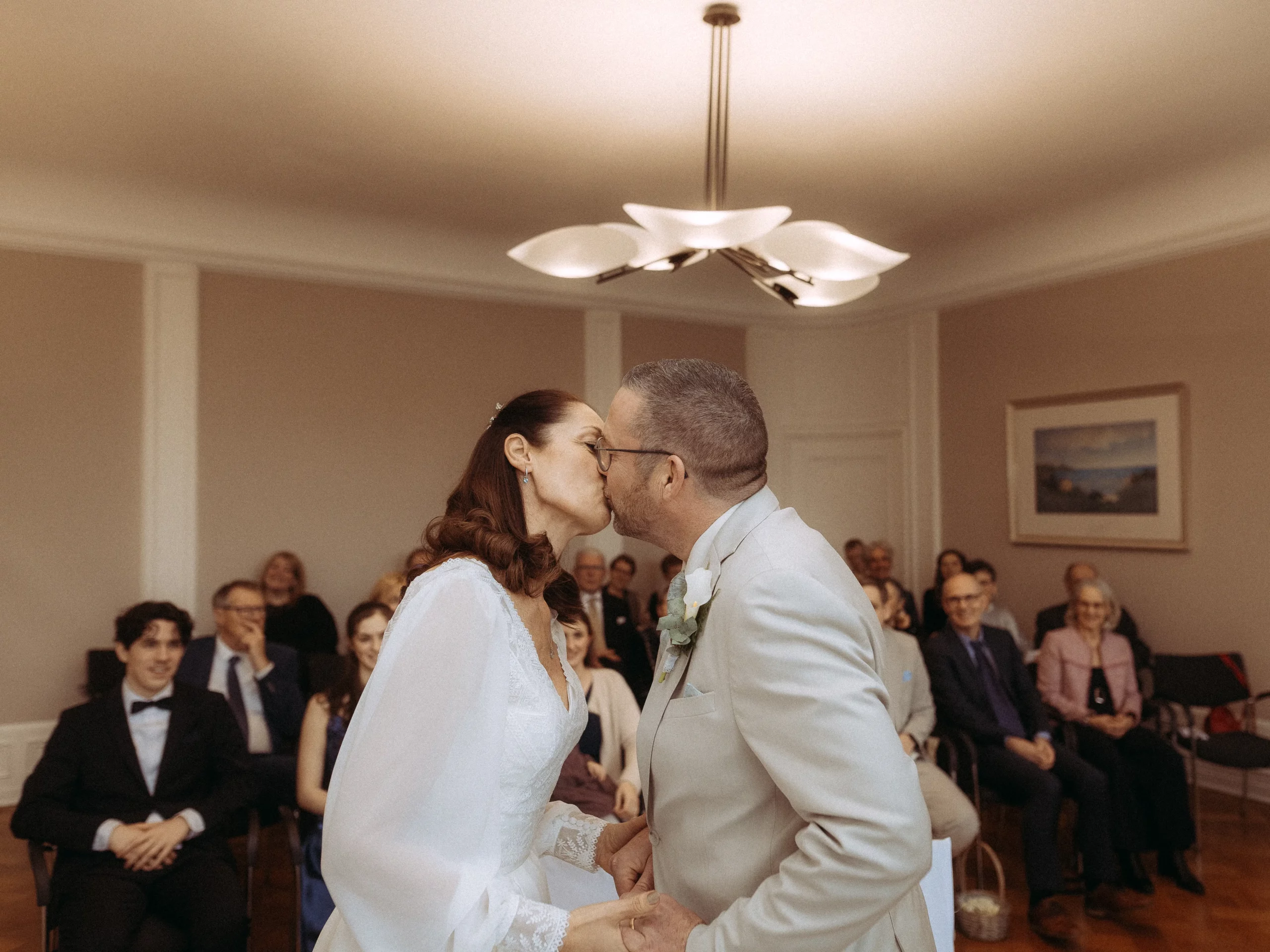 Ein Paar in Hochzeitskleidung küsst sich vor dem Altar in einem kleinen, warm beleuchteten Raum, während die Gäste lächeln. Wunderschön eingefangen von Hochzeitsfotograf MV.
