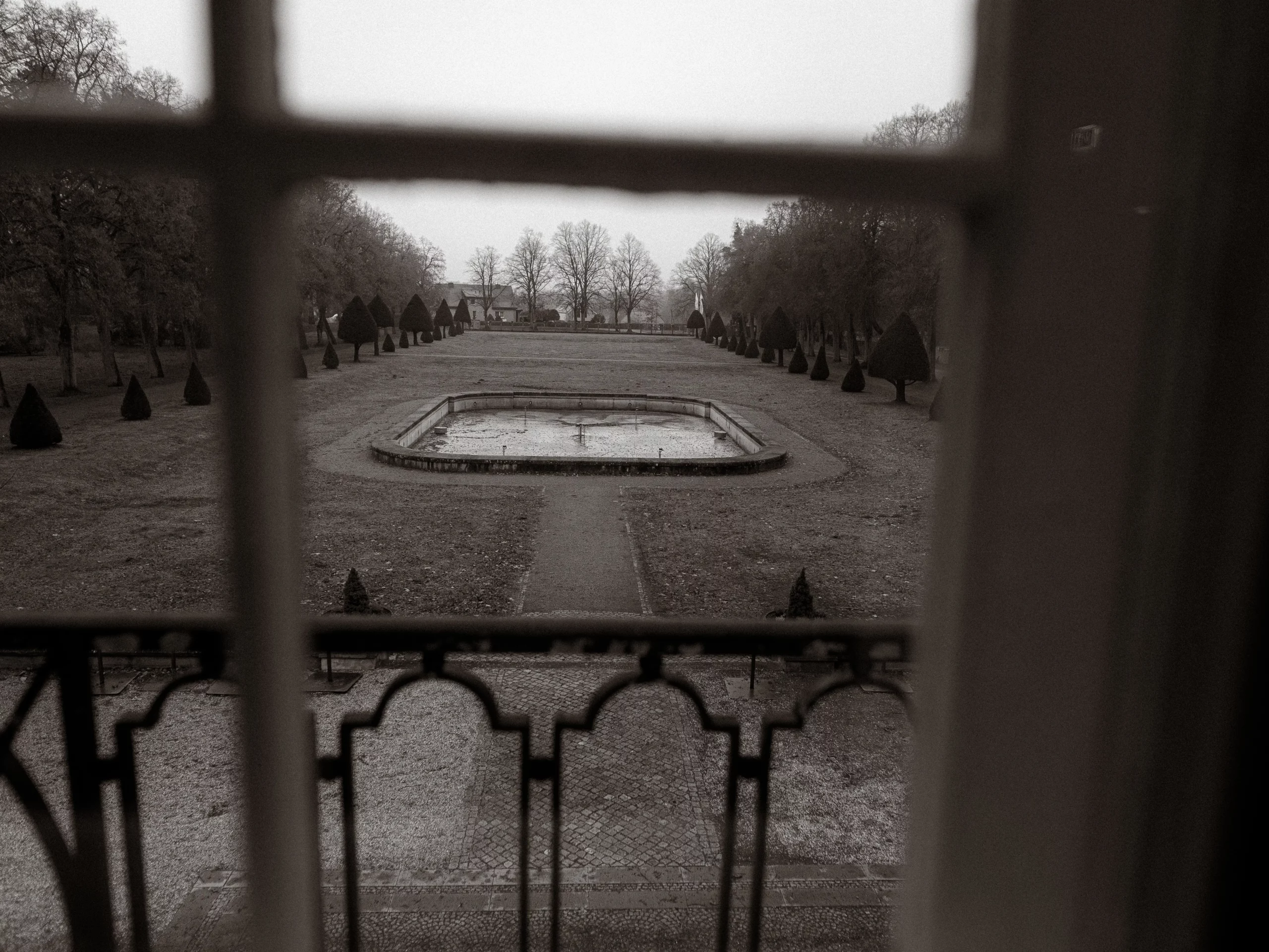 Ein Schwarz-Weiß-Foto, das durch ein Fenster aufgenommen wurde, zeigt einen trockenen, rechteckigen Brunnen in der Mitte eines formalen Gartens mit Bäumen und beschnittenen Büschen an einem bewölkten Tag - aufgenommen von Hochzeitsfotograf MV.