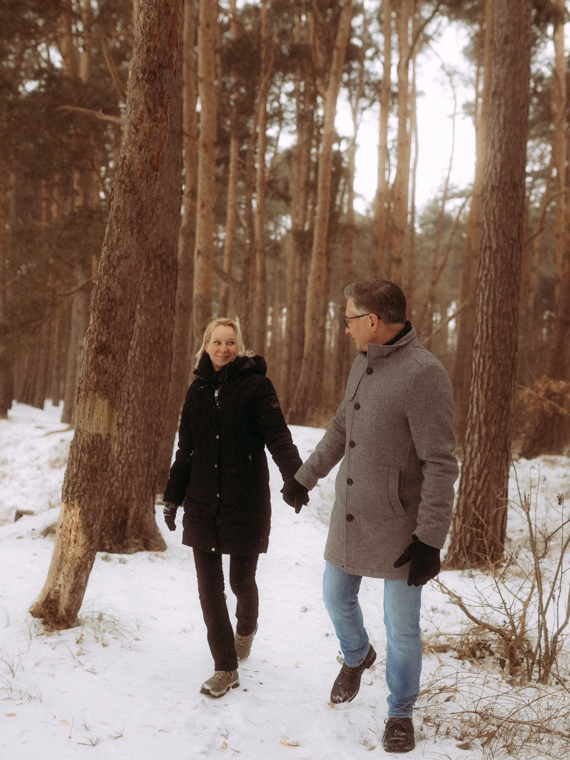 Ein Mann und eine Frau gehen Hand in Hand durch einen verschneiten Wald, in Wintermäntel und Handschuhe gehüllt, und lächeln sich zwischen hohen Bäumen an. Dieser von Fotograf MV aufgenommene Moment strahlt Wärme in einer winterlichen Landschaft aus.