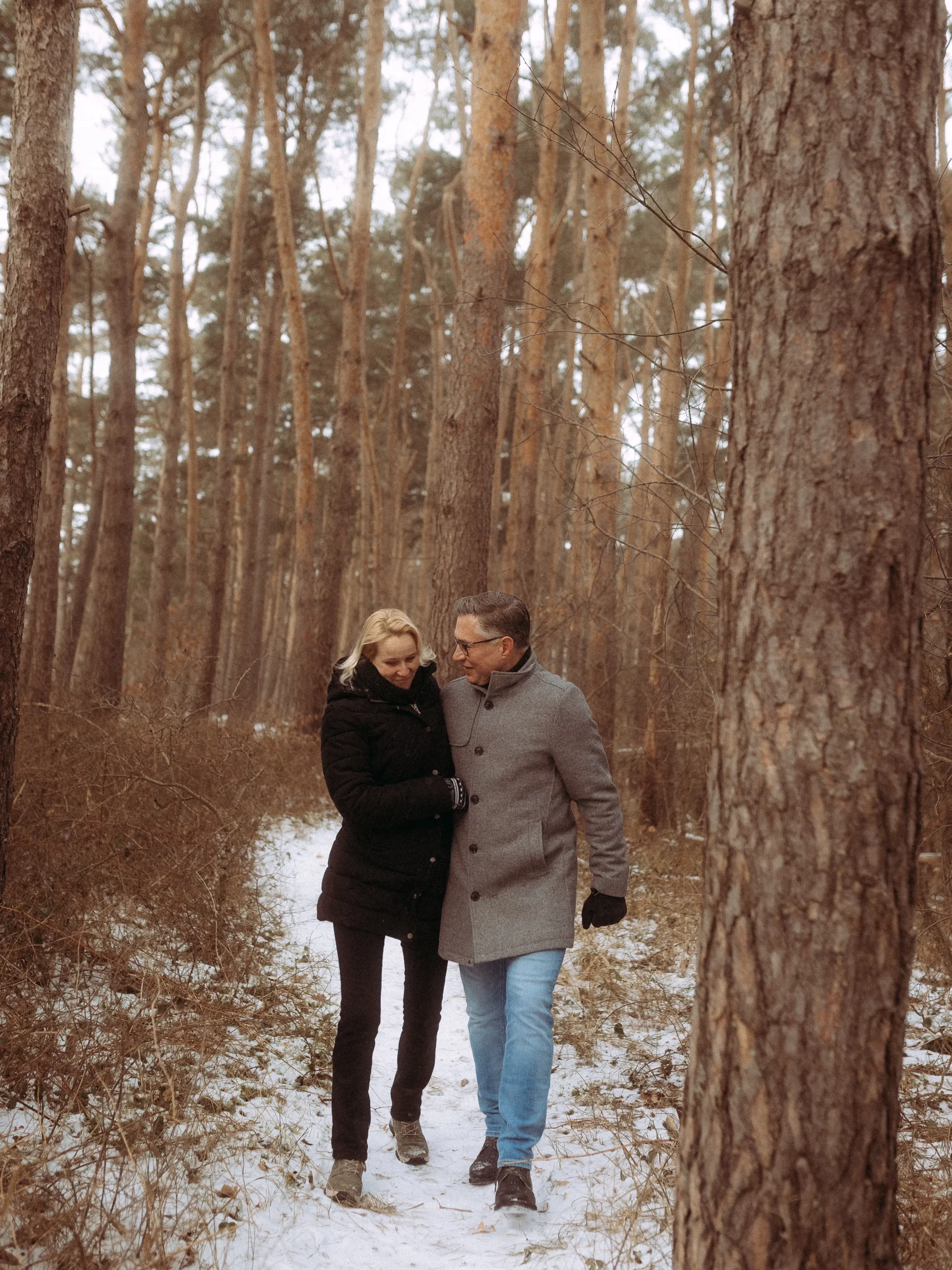 Ein Paar geht eng beieinander auf einem verschneiten Weg durch einen Wald mit hohen, dünnen Bäumen. Beide tragen Wintermäntel, während sie die ruhige, intime Atmosphäre genießen - perfekt, um besondere Momente mit einem Hochzeitsfotograf MV festzuhalten.