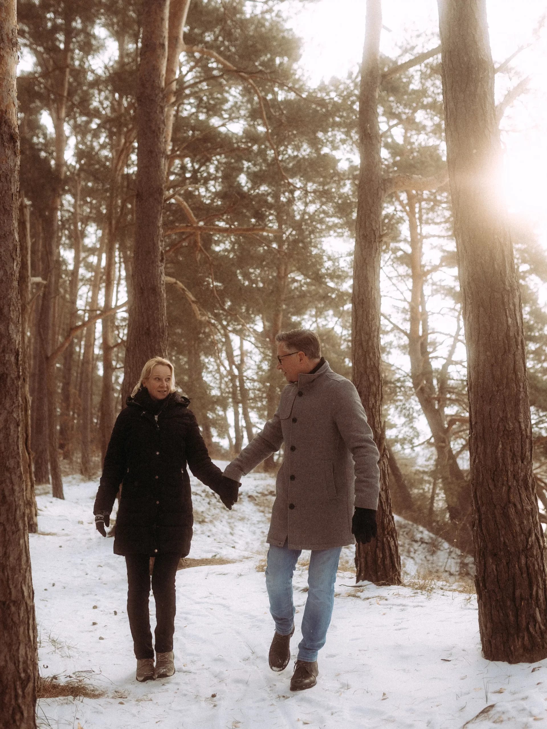 Ein Paar in Wintermänteln und Handschuhen geht Hand in Hand einen verschneiten Waldweg entlang, umgeben von hohen Bäumen, durch deren Äste das Sonnenlicht scheint - perfekt für Hochzeitsfotos MV.