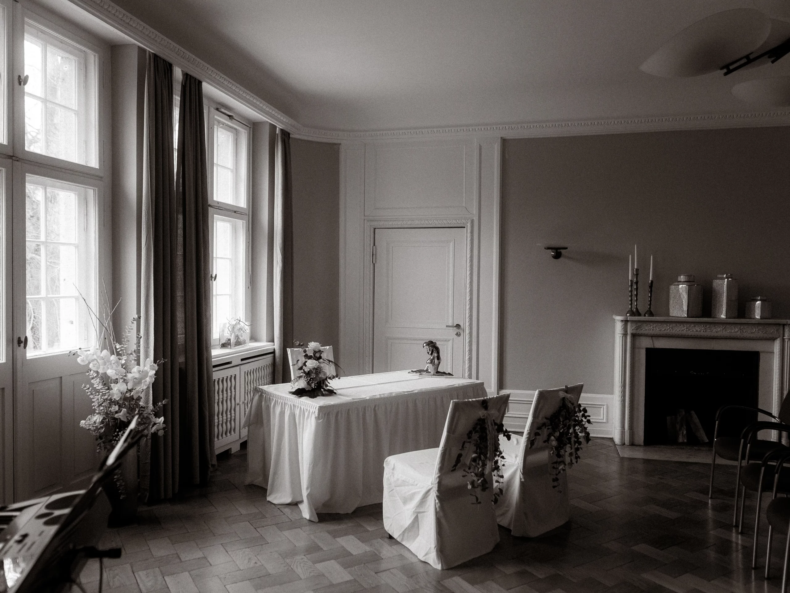 Ein Schwarz-Weiß-Foto von Fotograf MV zeigt einen minimalistisch eingerichteten Raum mit zwei Stühlen, einem mit einem weißen Tuch bedeckten Tisch, Blumenarrangements, großen Fenstern mit Vorhängen und einem mit Kerzen und Vasen geschmückten Kamin.