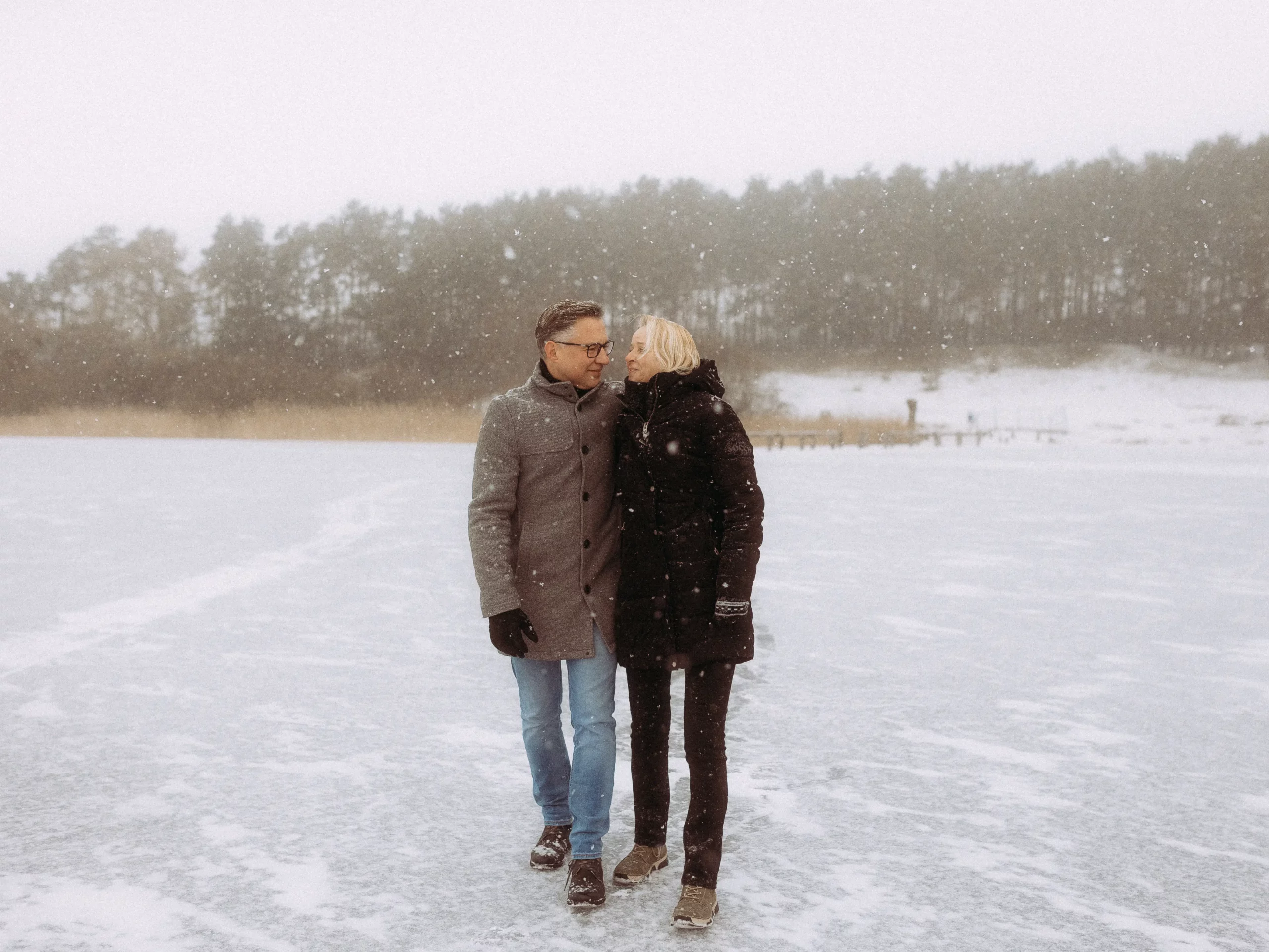 Ein Paar geht eng beieinander auf einem verschneiten, zugefrorenen See, umgeben von schneebedeckten Bäumen. Der Mann trägt einen grauen Mantel und Jeans, die Frau einen schwarzen Mantel und eine Hose, während um sie herum sanft der Schnee fällt.