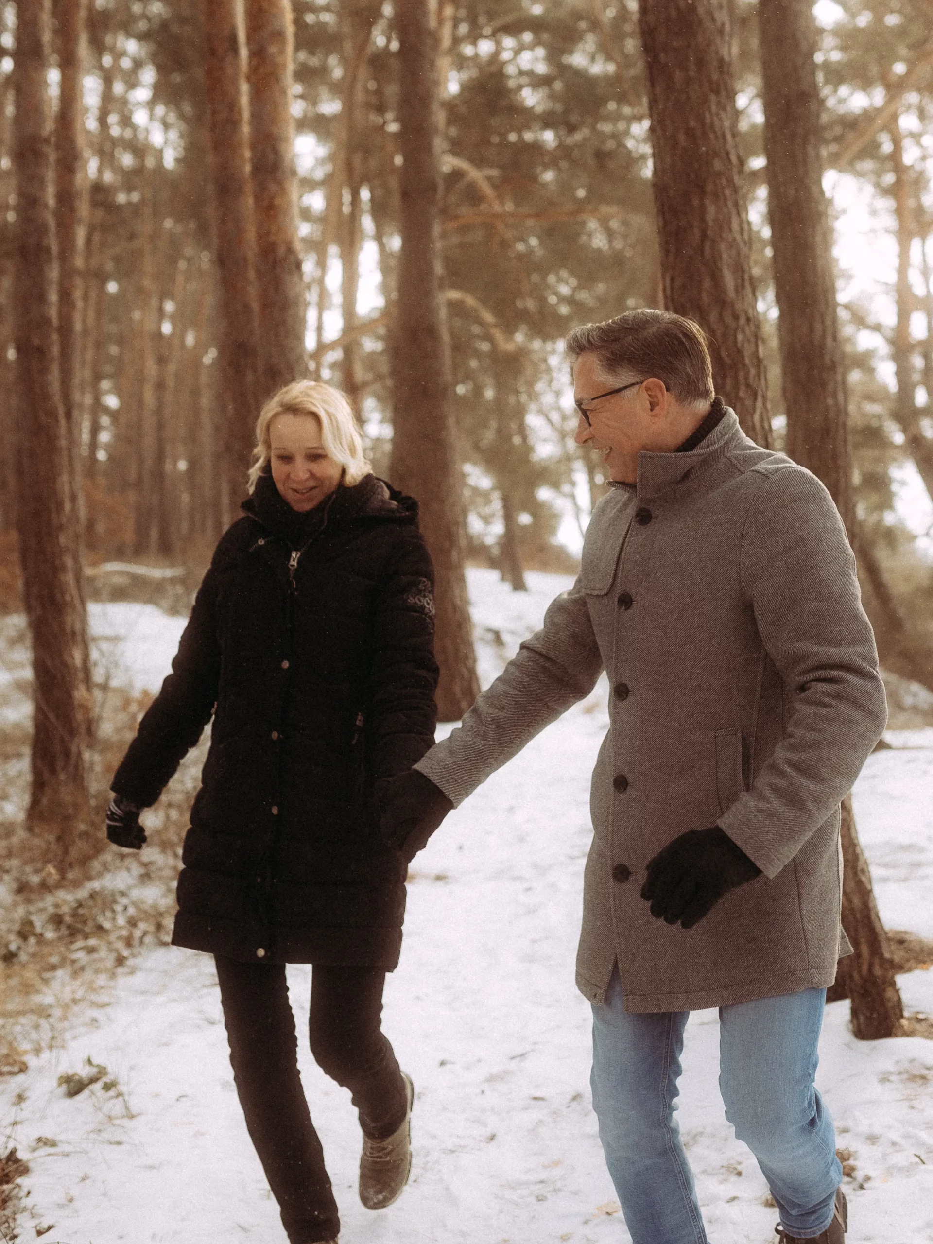 Ein Mann und eine Frau in Wintermänteln und Handschuhen spazieren Hand in Hand durch einen verschneiten Wald, lächelnd zwischen hohen Bäumen - wunderschön eingefangen von einem Familienfotografen MV.
