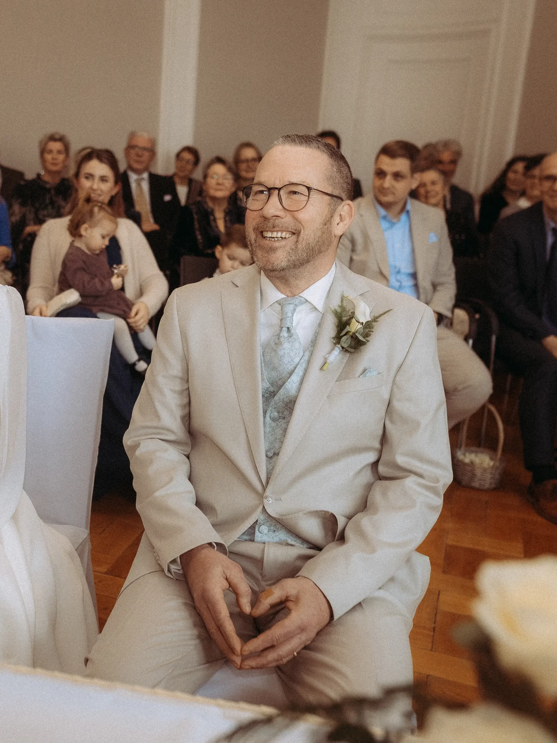 Ein Mann in einem beigen Anzug und blauer Krawatte sitzt lächelnd auf einer Hochzeitsfeier mit Gästen im Hintergrund. Wie von Hochzeitsfotograf MV festgehalten, trägt er eine Brille und eine Boutonnière am Revers.