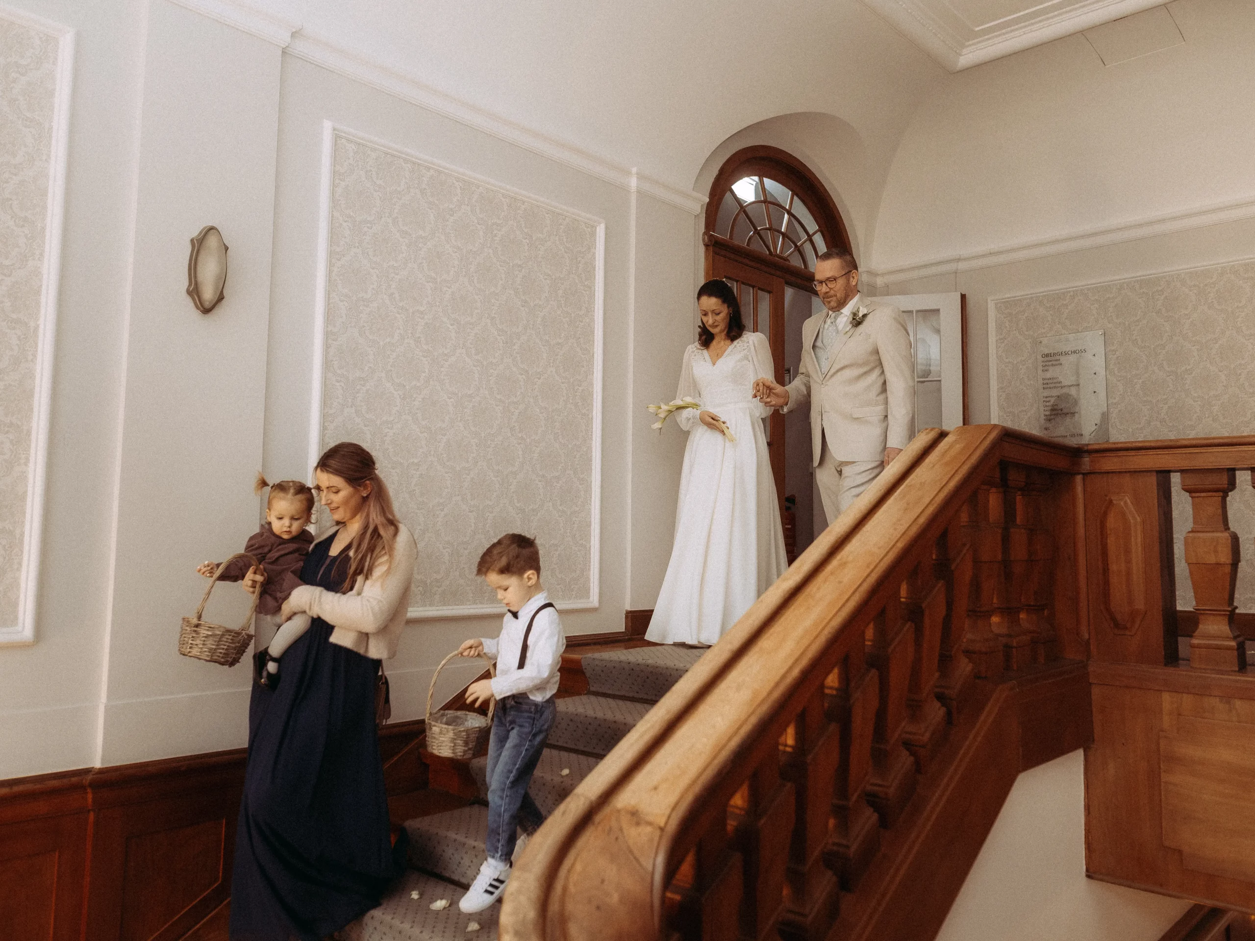 Braut und Bräutigam gehen händchenhaltend eine Treppe hinunter, gefolgt von einer Frau, die ein Kleinkind trägt, und einem kleinen Jungen mit einem Korb. Das elegante Interieur mit Holzgeländer ist von einem Hochzeitsfotograf aus Schwerin wunderschön eingefangen worden.