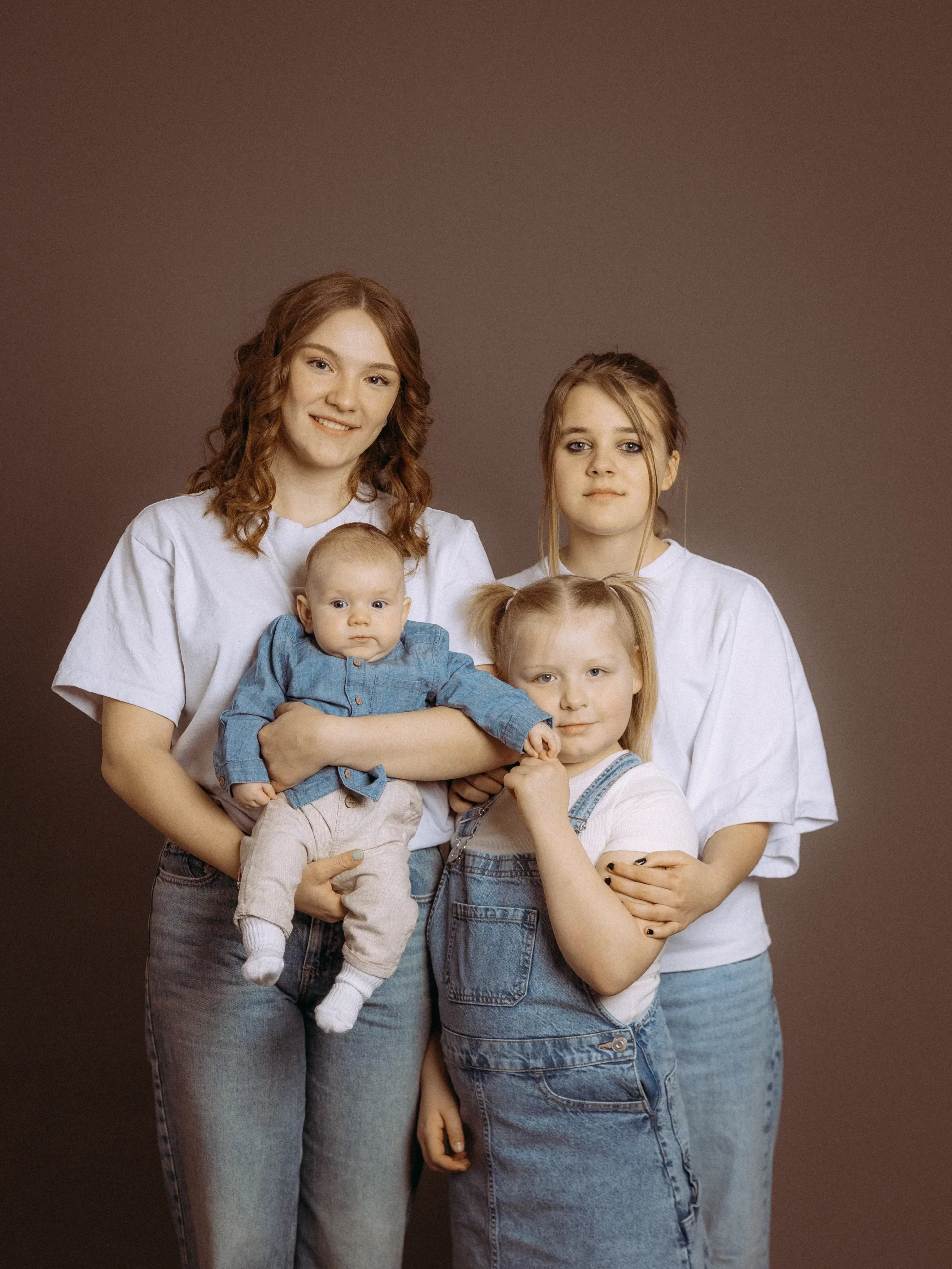 Familienfoto in MV: Zwei junge Frauen in Jeans und weißen TShirts halten ein Baby und ein kleines Mädchen mit blonden Zöpfen. Warme Farben, brauner Hintergrund. Fotograf aus Schwerin.