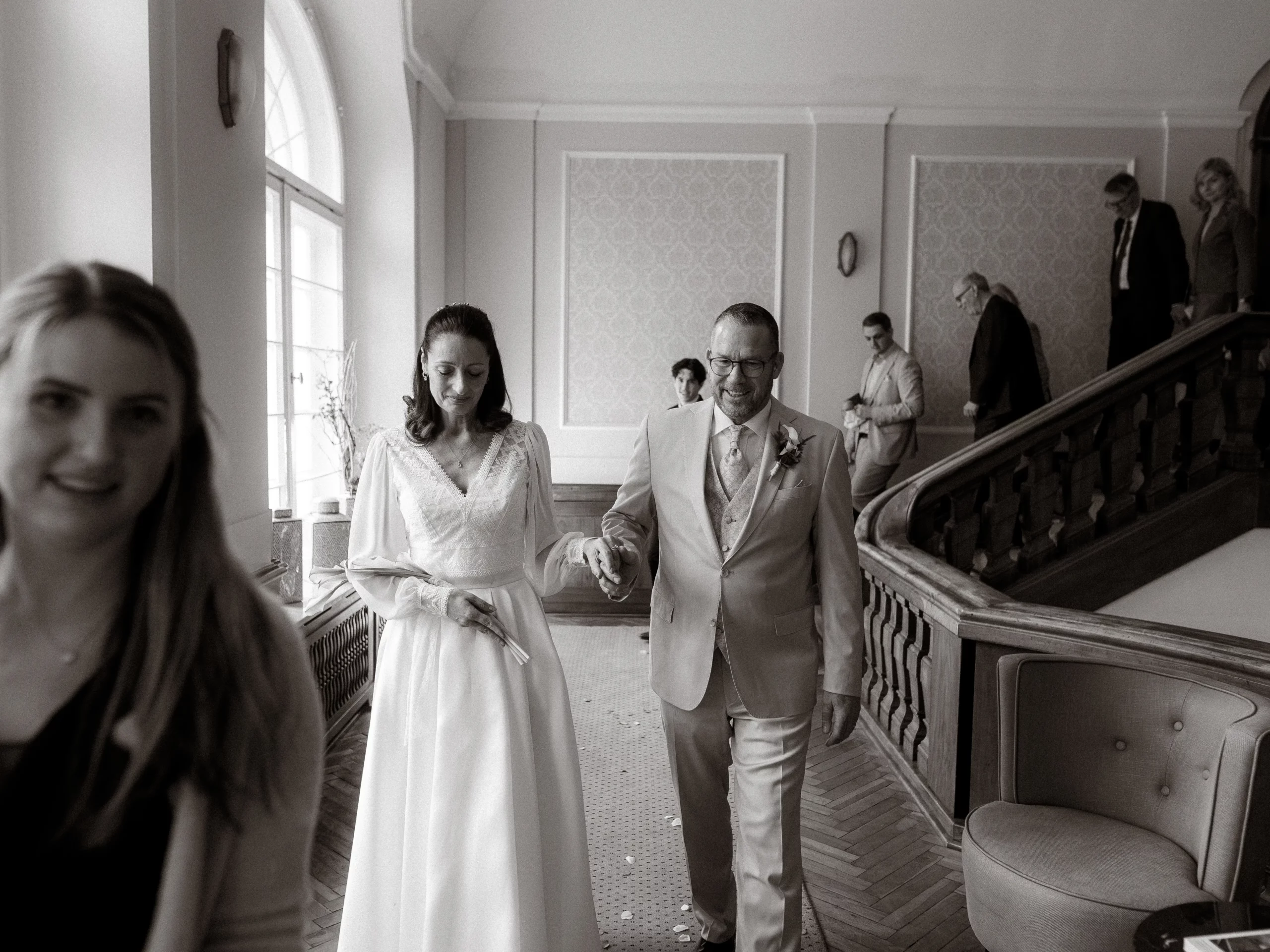 Eine Braut in einem weißen Kleid geht Hand in Hand mit einem lächelnden Mann im Anzug durch einen Flur, begleitet von anderen. Der Hochzeitsfotograf aus Schwerin hat diesen Moment in einer Hochzeitslocation aufgenommen, der Freude und Zweisamkeit ausstrahlt.
