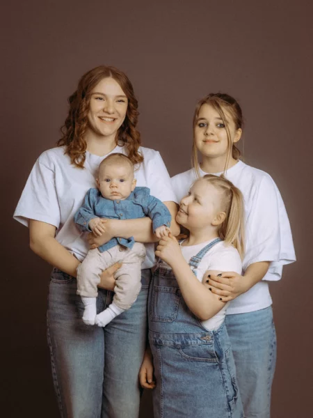 Familienfotos MV: Drei Schwestern umarmen sich und halten einen Säugling. Die älteste Schwester mit lockigen, roten Haaren lächelt. Die jüngste Schwester mit blonden Haaren schaut liebevoll zu ihr auf. Fotograf aus Schwerin.