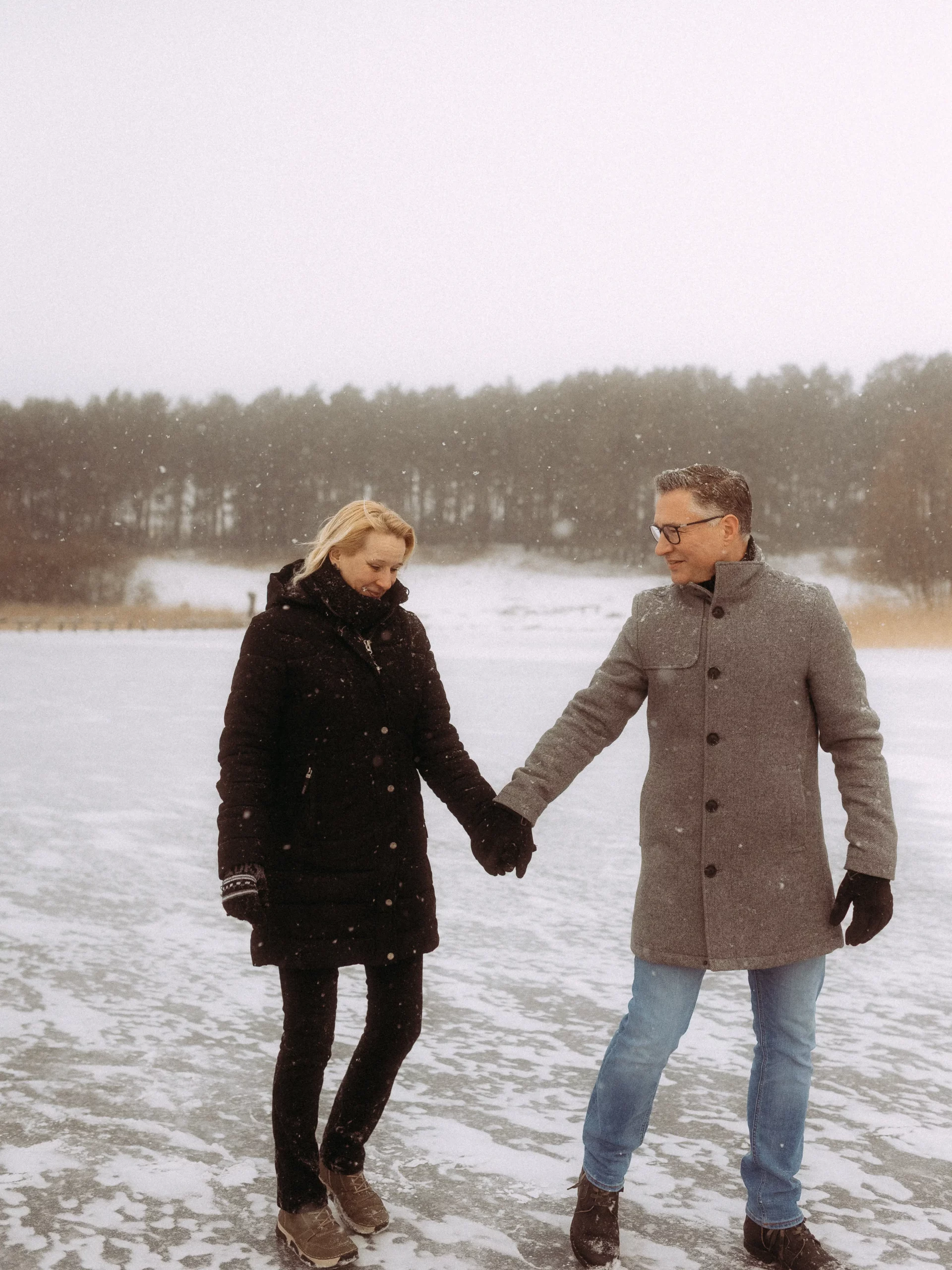Ein Paar hält sich auf einem zugefrorenen, schneebedeckten See an den Händen, gekleidet in Wintermantel und Handschuhe. Bäume und Schnee füllen den Hintergrund an einem bewölkten Tag - ein bezaubernder Moment, eingefangen von Hochzeitsfotograf MV.