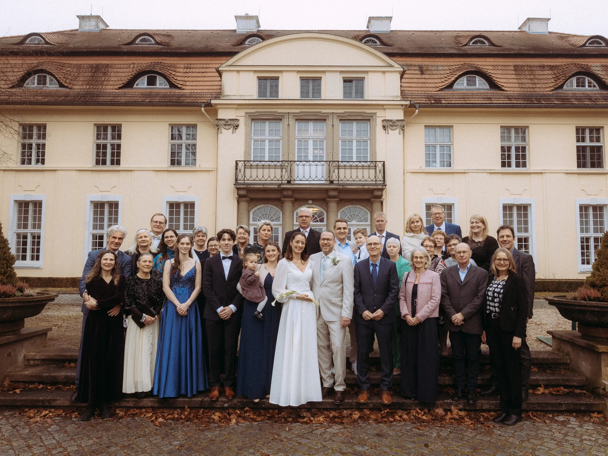 Eine große, förmlich gekleidete Gruppe posiert für ein Foto vor einem eleganten, historischen Herrenhaus mit hellgelben Wänden und hohen Fenstern. Im Mittelpunkt steht das Paar, das von einem Hochzeitsfotografen in MV bei seiner schönen Hochzeitsfeier festgehalten wird.