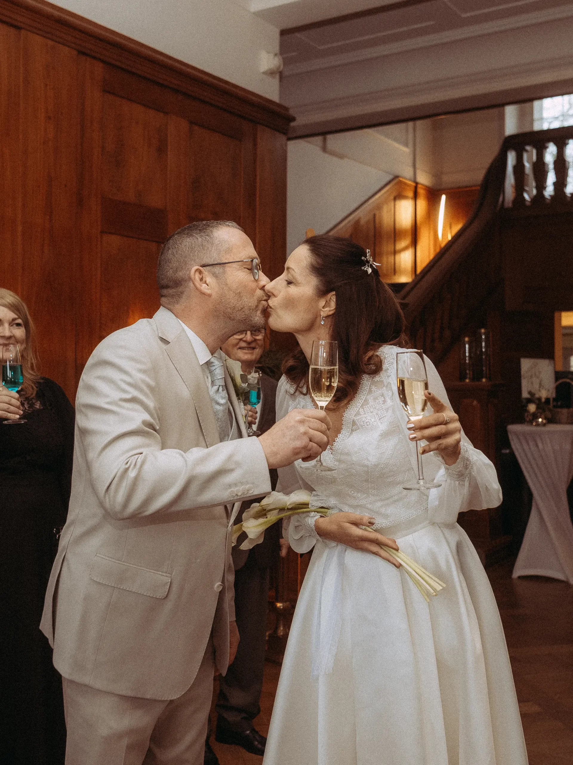 Ein weiß gekleidetes Brautpaar küsst sich mit Sektgläsern in der Hand auf ihrer Hochzeit, die in einem warm beleuchteten Raum mit Holzwänden und einer Treppe stattfindet - aufgenommen von einem Hochzeitsfotografen aus Schwerin.