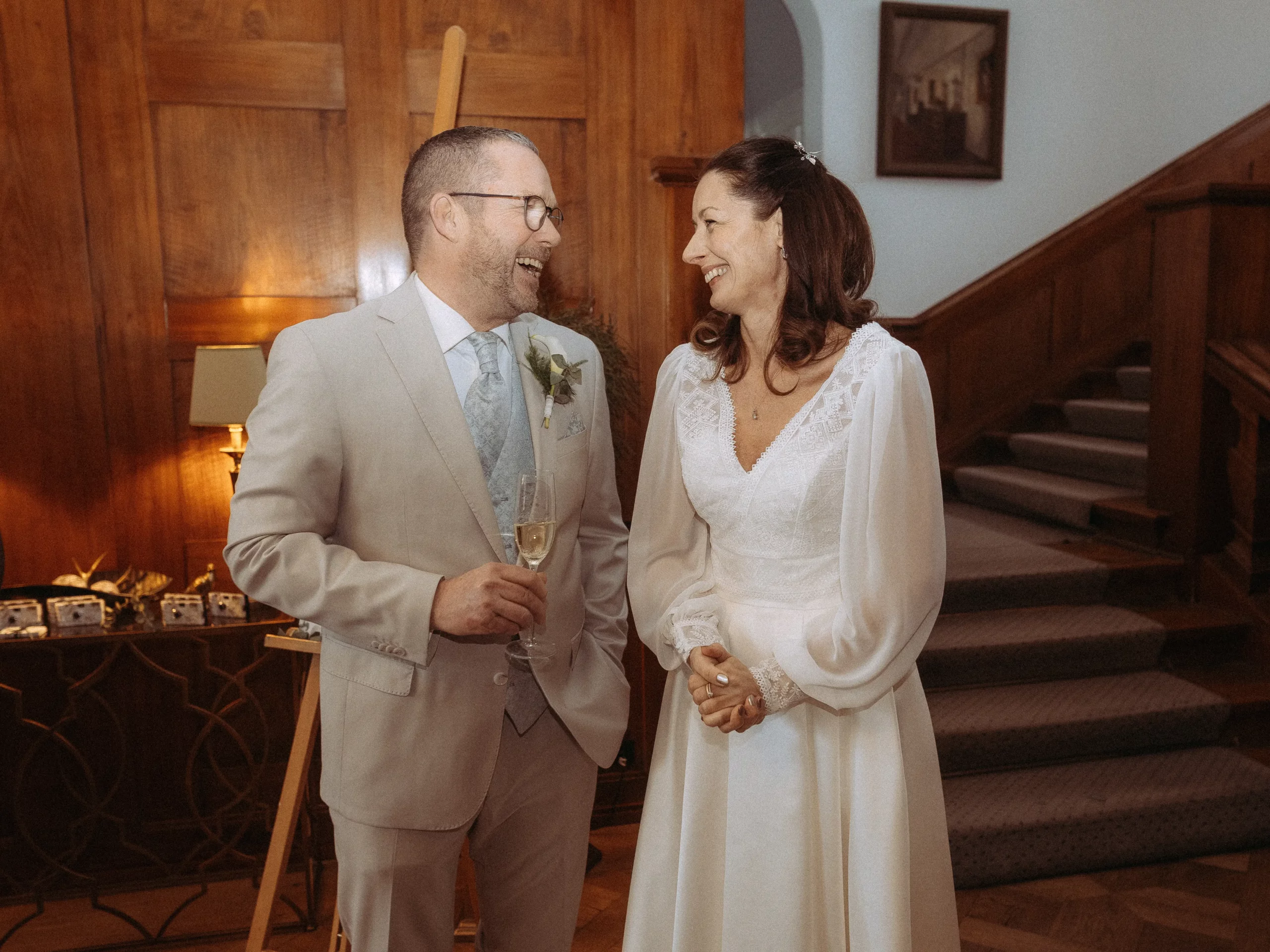 Ein lächelndes Paar in Hochzeitskleidung steht in einem Innenraum in der Nähe einer Treppe und lacht zusammen. Der Mann hält ein Sektglas in der Hand. Warme Beleuchtung und Holzvertäfelung setzen die Szene in Szene - perfekt für Hochzeitsfotos MV von einem erfahrenen Fotografen in MV.