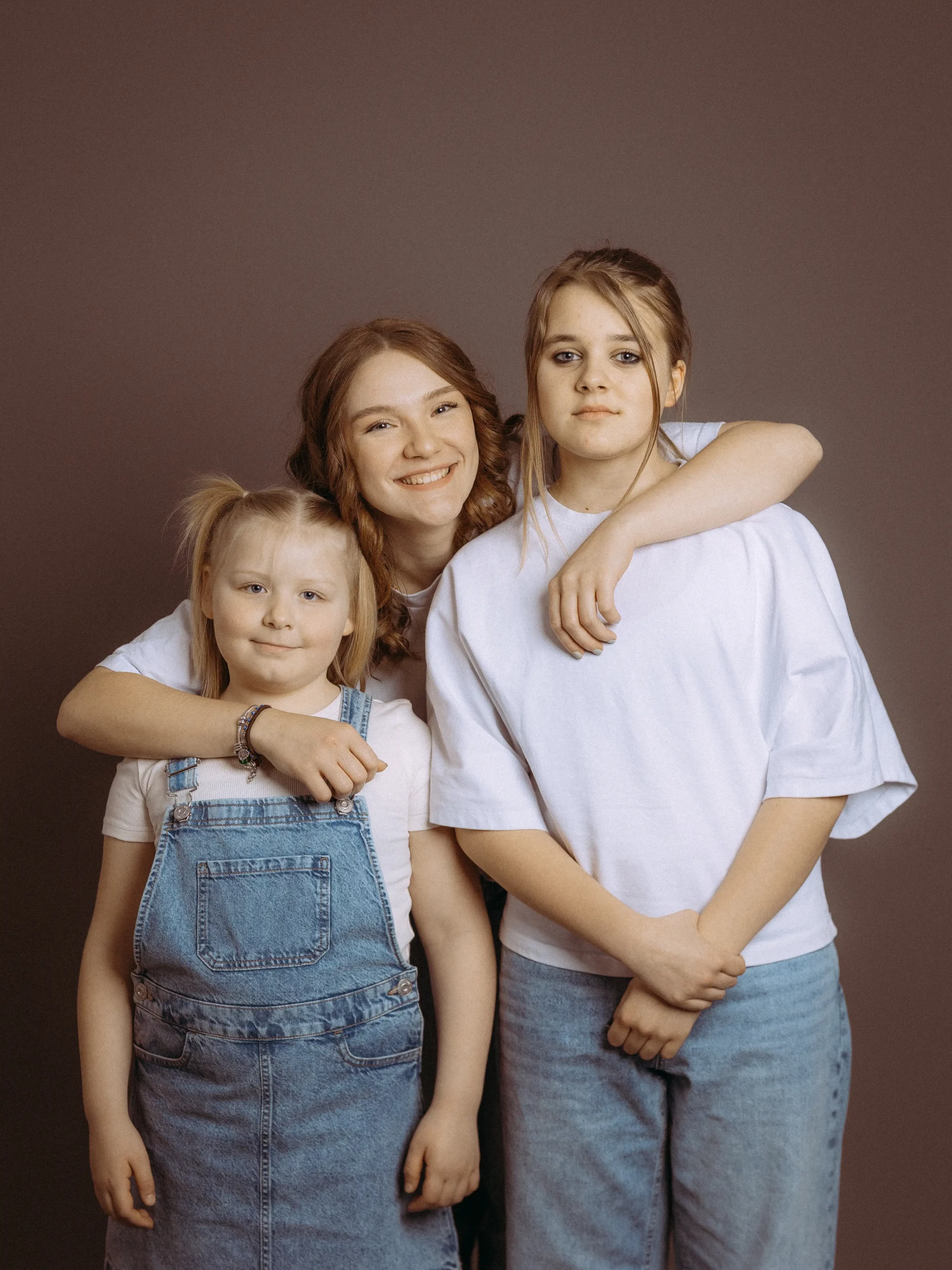Drei Schwestern, die jüngste in Jeanslatzhose, die älteren in Jeans und TShirts, posieren fröhlich. Familienfotos MV vom Fotograf aus Schwerin.