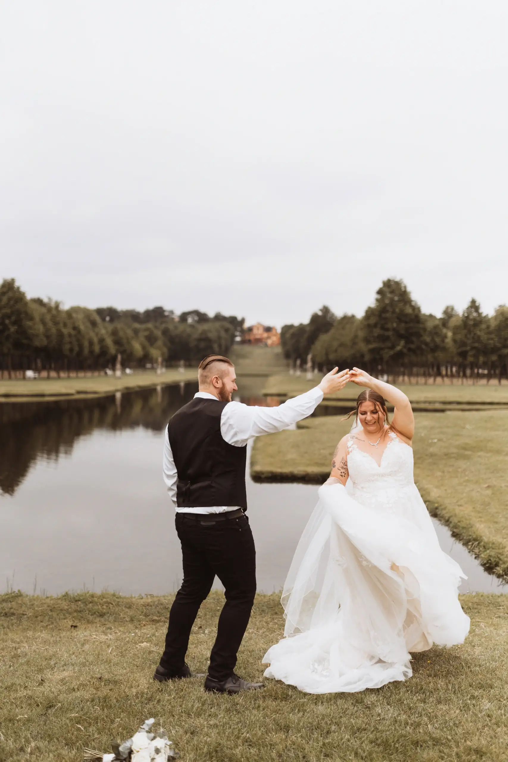 Ein glückliches Hochzeitspaar tanzt auf einer Wiese am See. Der Hochzeitsfotograf in MV hat den besonderen Moment eingefangen. Bäume und ein Schloss sind im Hintergrund zu sehen.