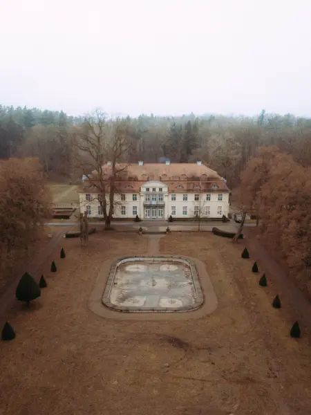 Ein elegantes, helles Herrenhaus im barocken Stil aus der Vogelperspektive, umgeben von einem herbstlichen Wald und einem leeren, rechteckigen Springbrunnen im Vordergrund. Die ruhige Atmosphäre macht das Anwesen zu einer idealen Location für einen Hochzeitsfotograf in Schwerin und MV.