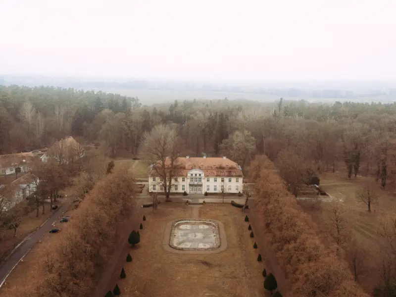 Drohnenaufnahme eines herrschaftlichen, hellen Herrenhauses mit rotem Ziegeldach, das von einem symmetrischen Garten mit kahlen Bäumen und einem leeren Brunnen umgeben ist. Die herbstliche Waldlandschaft im Hintergrund unterstreicht die ruhige Atmosphäre für einen Hochzeitsfotograf in Schwerin und MV.