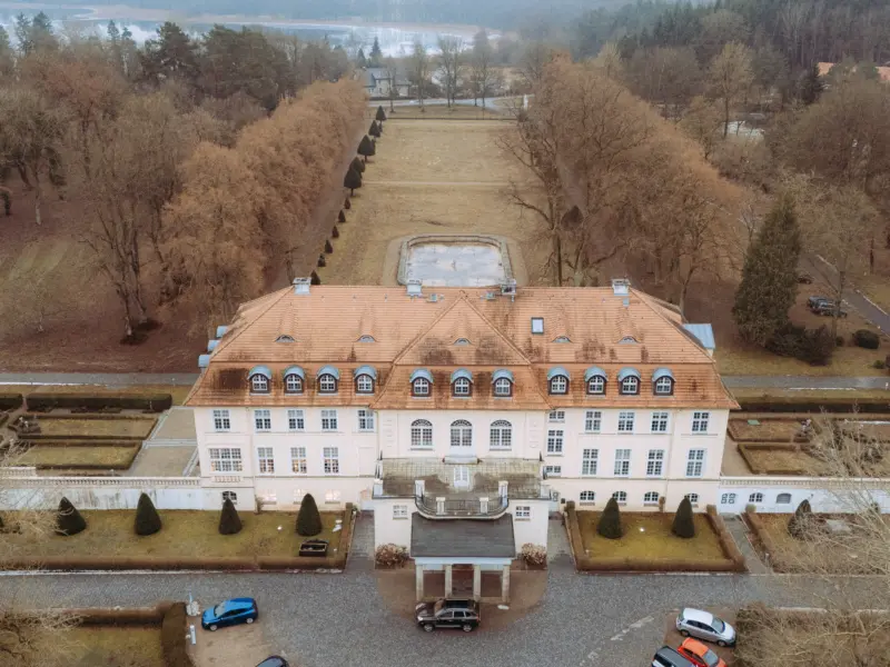 Drohnenaufnahme eines herrschaftlichen Schlosses mit sandfarbener Fassade und rotem Ziegeldach, umgeben von einem weitläufigen Park mit geometrischen Gartenanlagen und kahl gewordenen Bäumen. Ein idealer Ort für eine Hochzeit, festgehalten von einem Hochzeitsfotograf in Schwerin und MV.