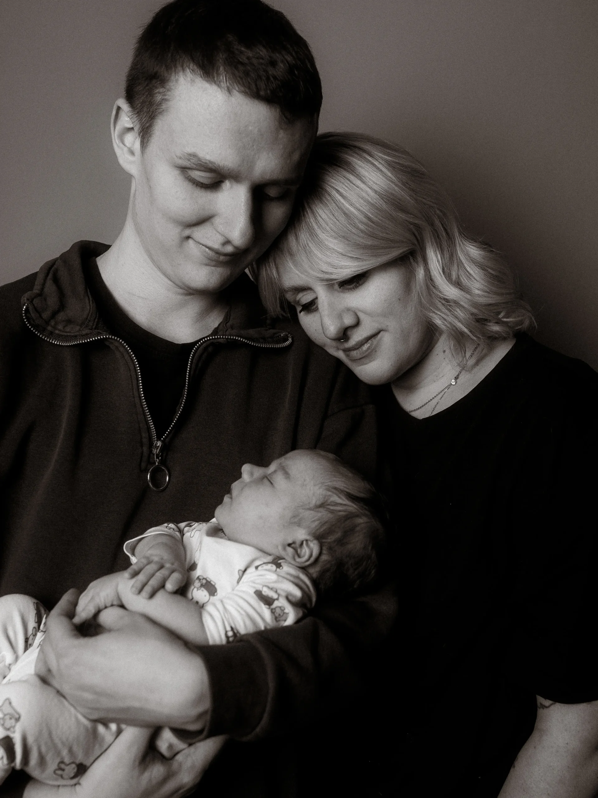 Ein zärtliches Porträt junger Eltern mit ihrem schlafenden Neugeborenen im Arm. Die Mutter und der Vater blicken liebevoll auf ihr Baby herab. Diese einfühlsame Aufnahme entstand als Familienfotograf in Schwerin in einem professionellen Fotostudio in Schwerin.