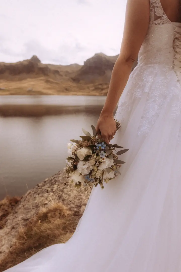 Eine Braut in einem eleganten weißen Spitzenkleid hält einen zarten Blumenstrauß vor einer malerischen Bergkulisse an einem ruhigen See. Die Szene fängt die romantische Stimmung perfekt ein – festgehalten von Ihrem kinematischen Hochzeitsfotograf in der Schweiz.