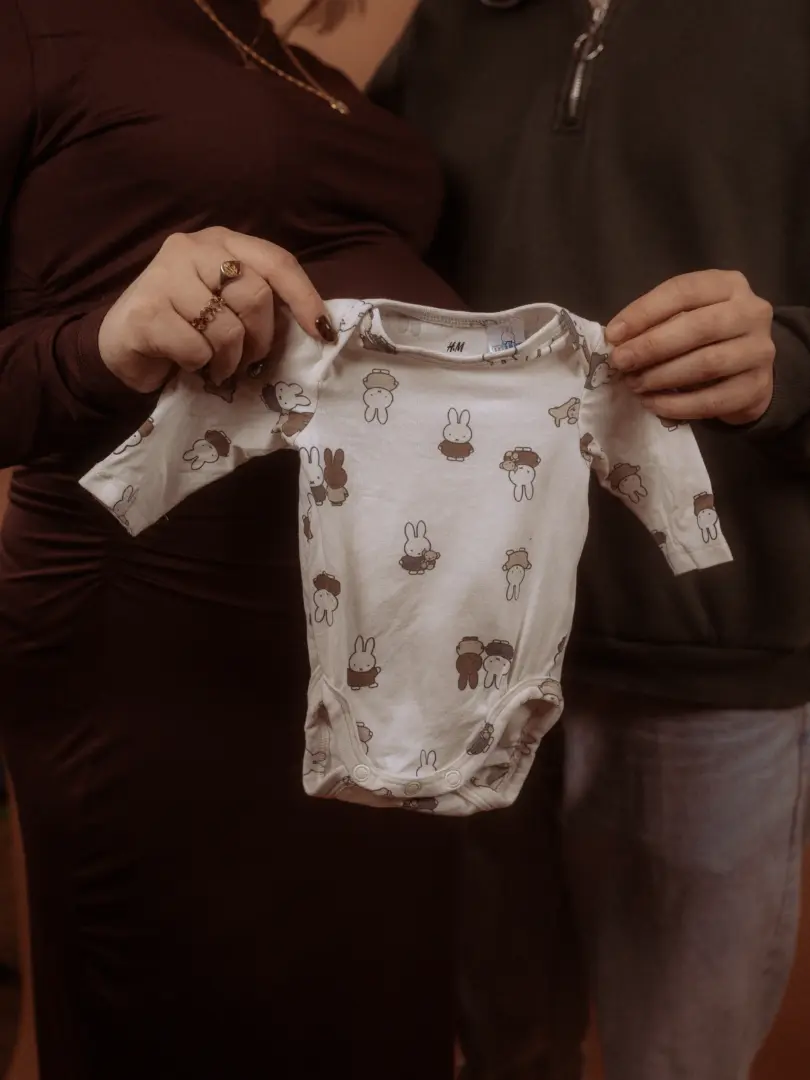 Ein Paar hält gemeinsam einen weißen Baby-Body mit Miffy-Hasen-Print vor den Babybauch der werdenden Mutter. Die Szene fängt eine emotionale Schwangerschaftsankündigung ein, ideal für Pärchenfotos in Schwerin.