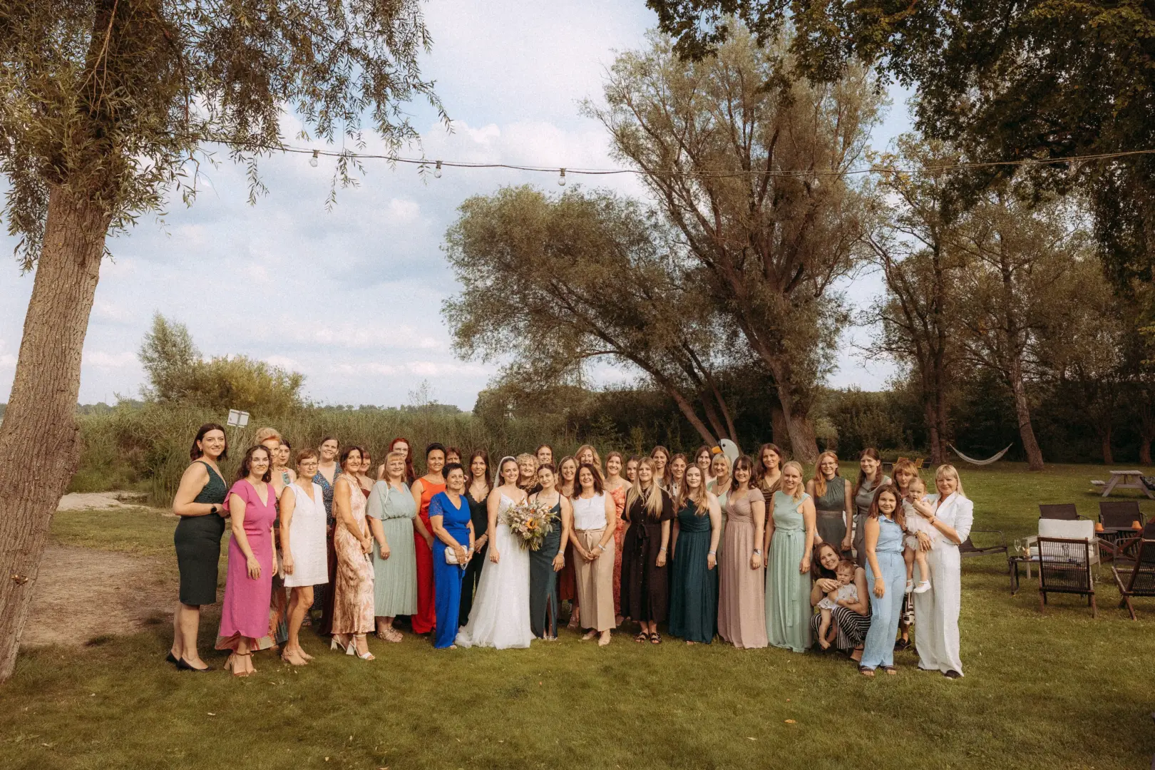 Eine große Hochzeitsgesellschaft mit der Braut in der Mitte posiert auf einer grünen Wiese vor einer malerischen Baumkulisse. Die Gäste tragen festliche Sommerkleider in verschiedenen Farben. Ein professioneller Hochzeitsfotograf in Schwerin und MV hat diesen Moment festgehalten.