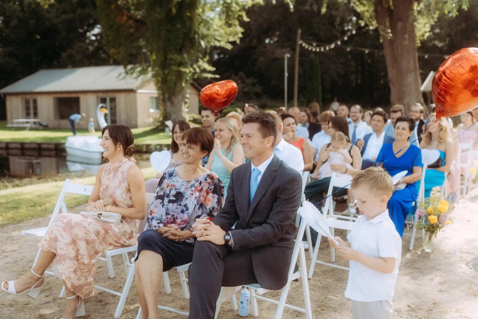 Gäste bei einer entspannten Outdoor-Trauung im Grünen, festgehalten vom Hochzeitsfotograf in Schwerin und MV. Eine Frau im Blumenkleid, ein Mann im Anzug und ein kleiner Junge mit einem weißen Fächer sitzen in der ersten Reihe, während im Hintergrund weitere Hochzeitsgäste zusehen.