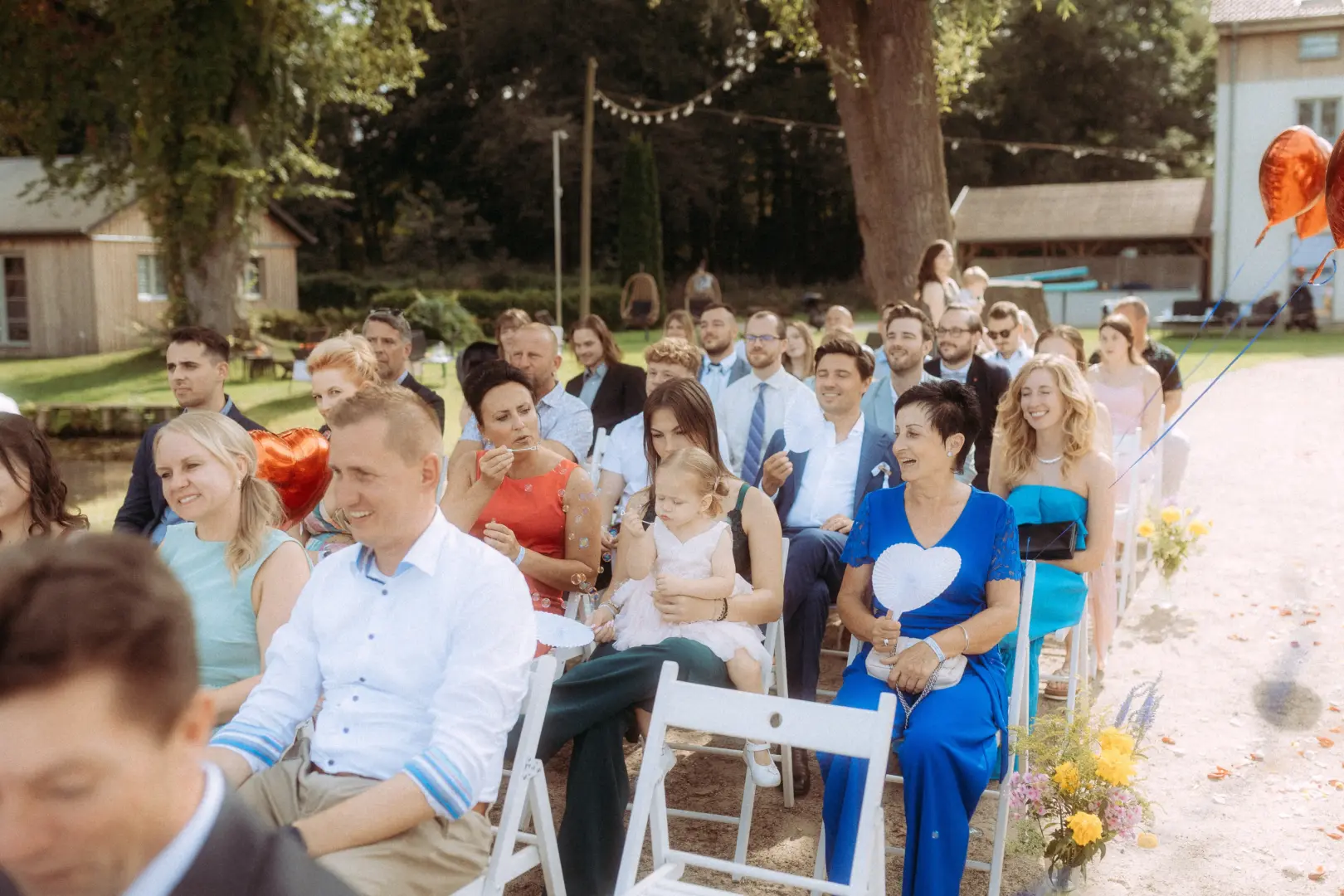 Gäste bei einer sommerlichen Outdoor-Trauung, bei der eine Frau in einem leuchtend blauen Kleid mit einem Fächer sitzt, während eine Mutter ihr kleines Kind auf dem Schoß hält. Die Stimmung ist festlich und entspannt, festgehalten von einem Hochzeitsfotograf in Schwerin und MV.