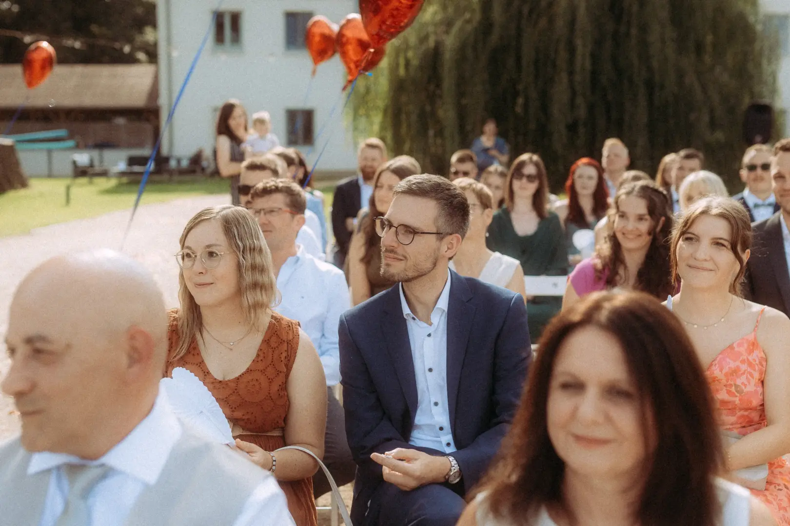 Gäste bei einer feierlichen Trauung im Freien, aufgenommen von einem Hochzeitsfotograf in Schwerin und MV. Die Hochzeitsgesellschaft sitzt entspannt in Reihen, während im Hintergrund rote Herzballons in den Himmel steigen und eine sommerliche, festliche Atmosphäre unterstreichen.