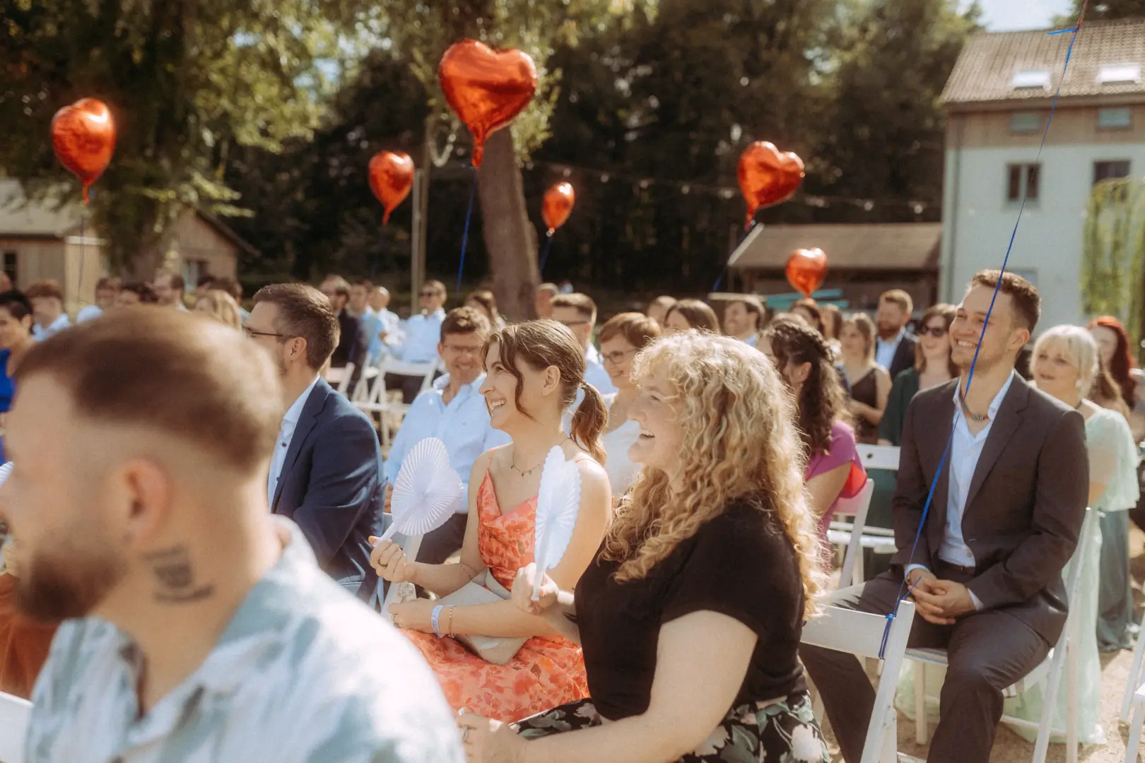 Gäste bei einer sommerlichen Outdoor-Trauung, die lachend in die Kamera blicken, während rote herzförmige Luftballons im Hintergrund schweben. Die festliche Stimmung unterstreicht die Arbeit als Hochzeitsfotograf in Schwerin und MV bei dieser emotionalen Zeremonie.