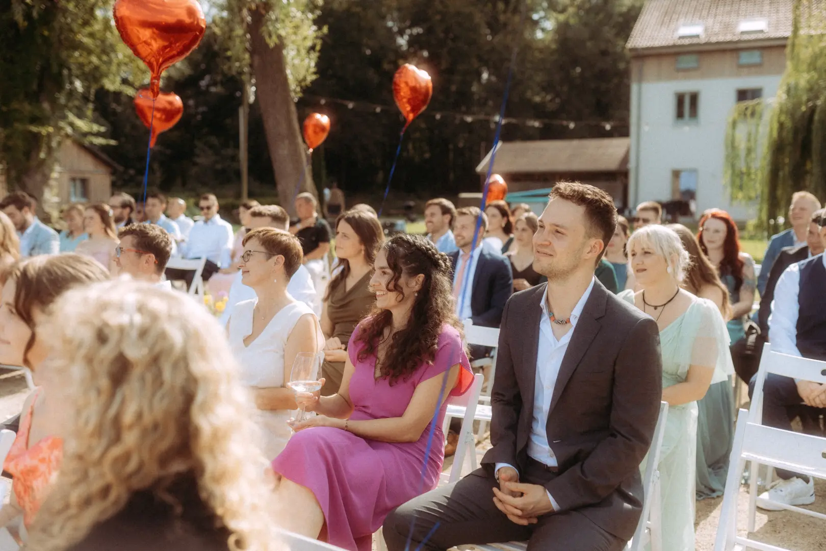 Gäste bei einer sommerlichen Trauung unter freiem Himmel mit roten Herz-Luftballons. Im Vordergrund sitzt ein glückliches Paar, während weitere Hochzeitsgäste gespannt zuschauen. Professioneller Hochzeitsfotograf in Schwerin und MV für authentische Hochzeitsreportagen.