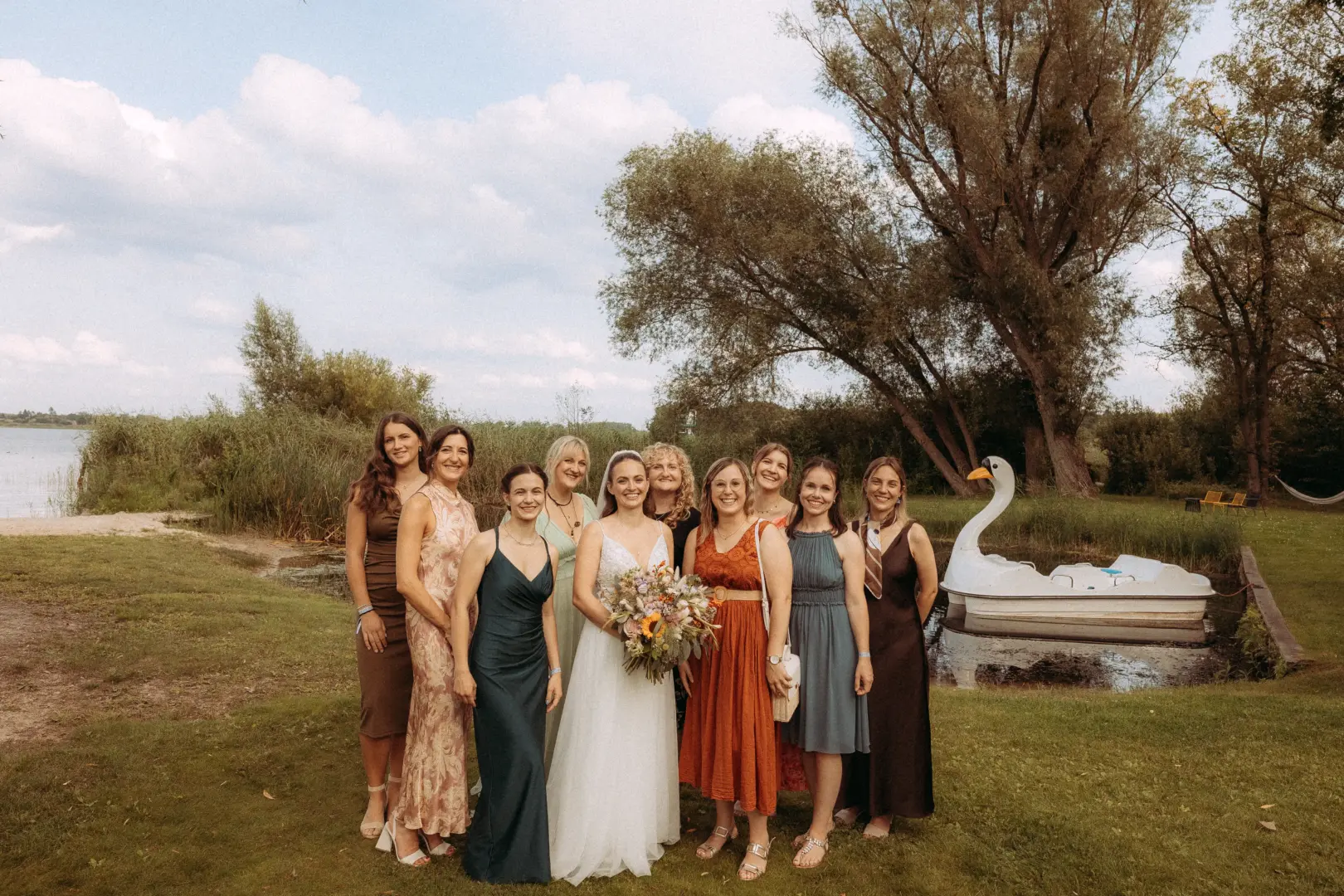 Eine Braut im weißen Hochzeitskleid mit Blumenstrauß posiert gemeinsam mit ihren Brautjungfern in verschiedenen eleganten Kleidern auf einer grünen Wiese am See. Im Hintergrund schwimmt ein Schwanen-Tretboot, fotografiert von einem Hochzeitsfotograf Müritz und MV.