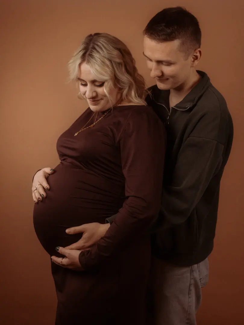 Ein werdendes Elternpaar bei einem professionellen Shooting im Fotostudio in Schwerin. Die Frau trägt ein dunkelrotes Kleid und hält ihren Babybauch, während ihr Partner liebevoll hinter ihr steht. Die warme, einfarbige Kulisse unterstreicht die intime Atmosphäre dieser Pärchenfotos.