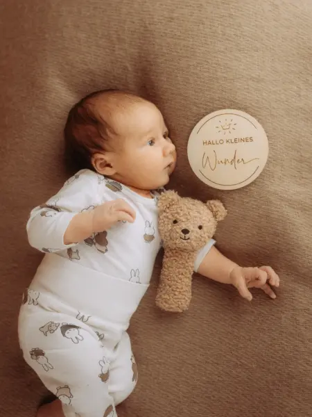 Ein neugeborenes Baby liegt entspannt auf einer beigefarbenen Decke, bekleidet mit einem weißen Strampler mit Hasenmuster. Neben dem Baby liegt ein kleiner Teddybär und eine hölzerne Erinnerungstafel mit der Aufschrift „Hallo kleines Wunder“. Familienfotograf in Schwerin.