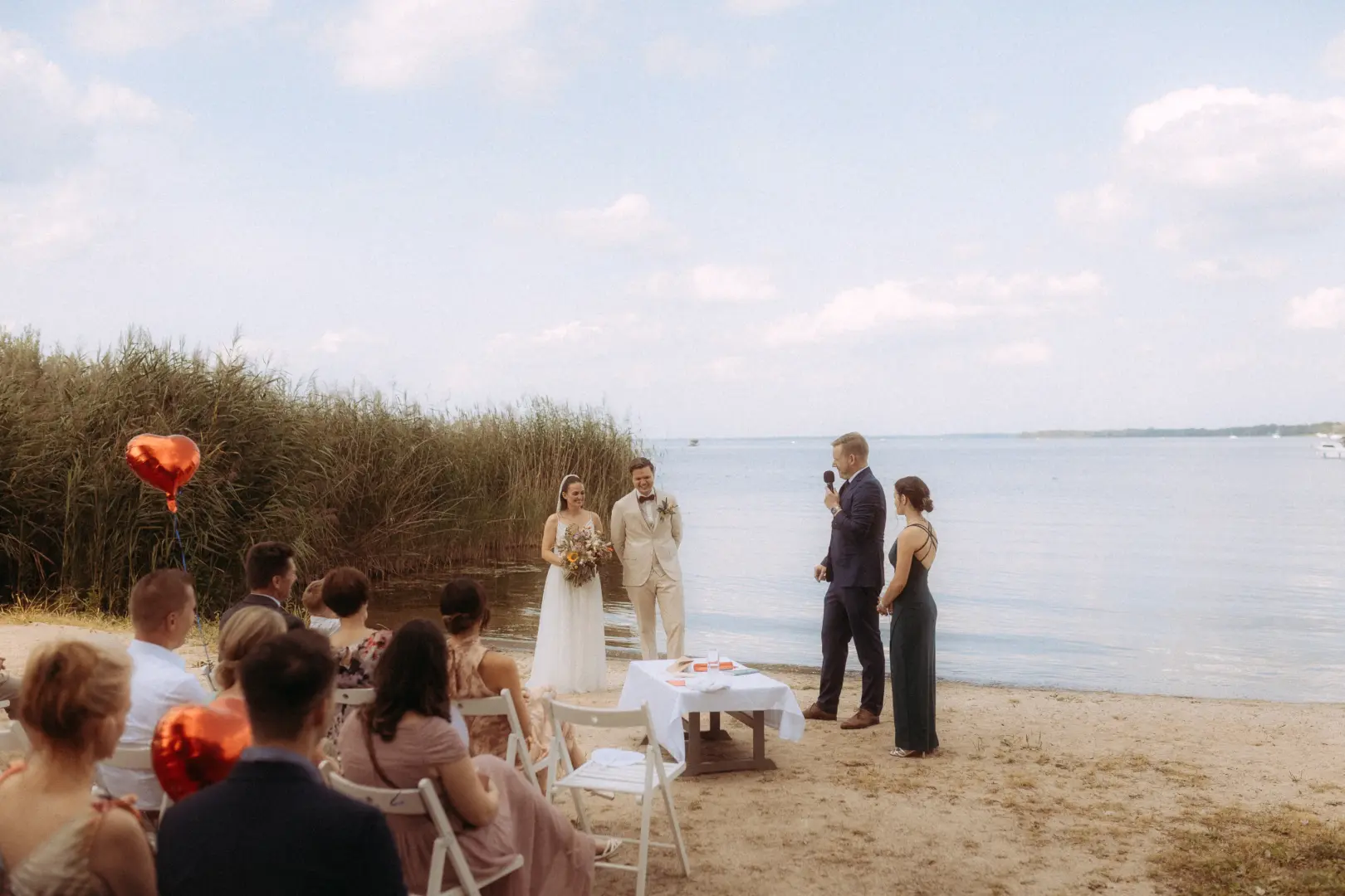 Ein Brautpaar steht während einer Zeremonie am sonnigen Strand eines Sees, während ein Redner das Wort ergreift. Die Szene fängt die emotionale Atmosphäre ein, die ein professioneller Hochzeitsfotograf in Schwerin und MV bei einer freien Trauung am Wasser festhält.