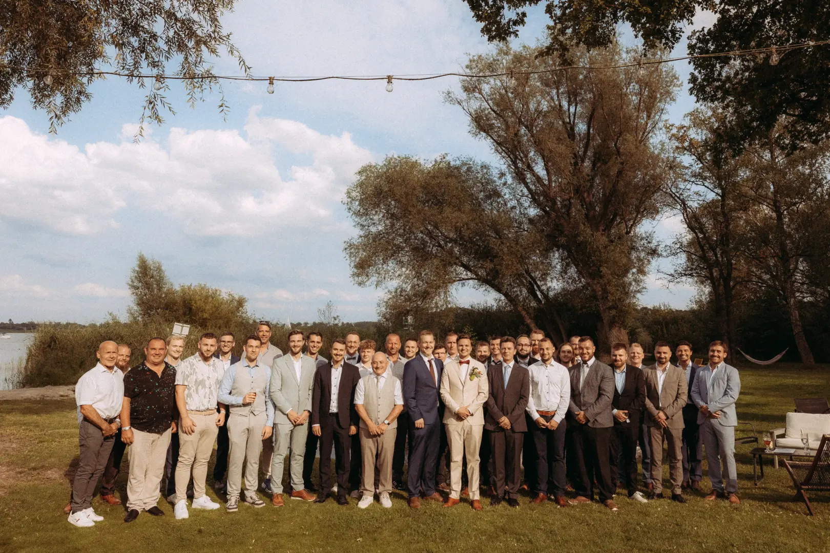 Eine große Gruppe gut gekleideter Männer posiert lächelnd für ein Foto im Freien bei einer Hochzeitsfeier. Die Gäste tragen Anzüge und sommerliche Outfits in einer grünen Parklandschaft am Wasser. Hochzeitsfotograf in Schwerin und MV.