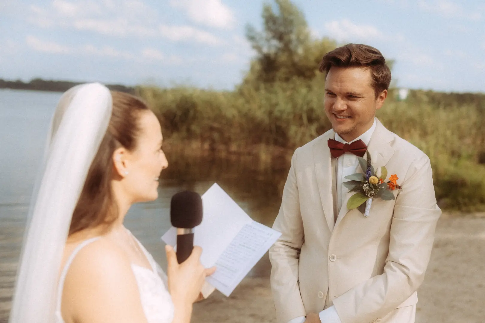Eine Braut trägt ihr Eheversprechen bei einer Zeremonie am See vor, während der Bräutigam in einem beigen Anzug mit weinroter Fliege sie liebevoll ansieht. Ein emotionaler Moment festgehalten von deinem Hochzeitsfotograf in Schwerin und MV.