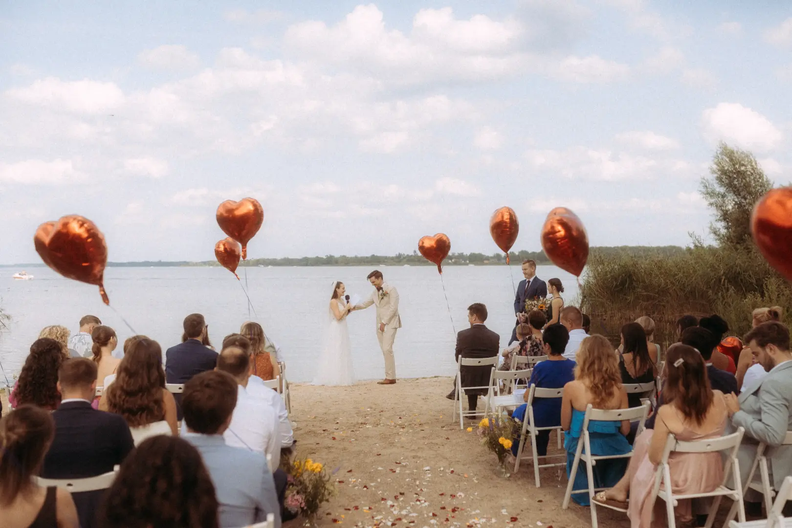 Ein Brautpaar tauscht während einer romantischen Zeremonie am Seeufer das Eheversprechen aus, umrahmt von herzförmigen Luftballons. Gäste sitzen auf weißen Stühlen im Sand. Professionelle Hochzeitsfotografie durch Ihren Hochzeitsfotograf Müritz und MV für unvergessliche Momente.