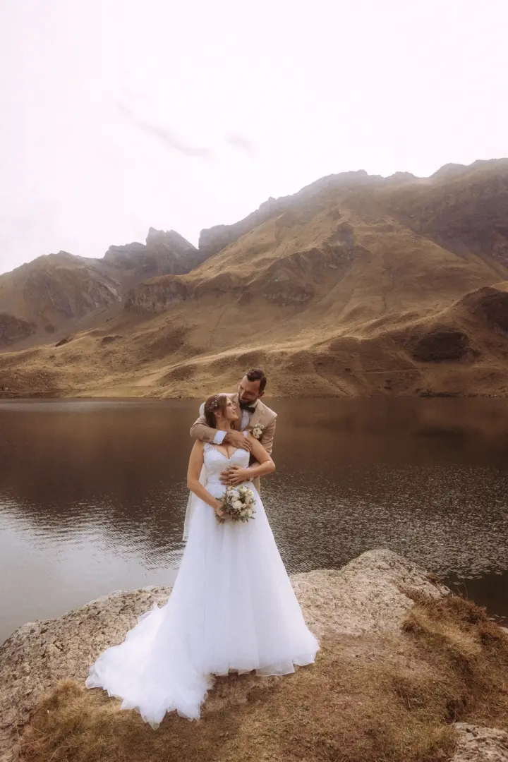 Ein Brautpaar in romantischer Umarmung an einem ruhigen Bergsee vor einer imposanten Gebirgskulisse – ein stimmungsvolles Motiv von einem kinematischen Hochzeitsfotograf in der Schweiz.