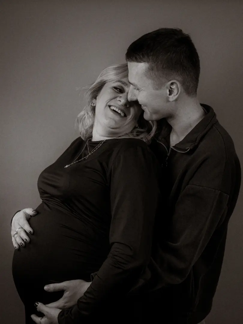 Ein glückliches schwangeres Paar in einem Fotostudio in Schwerin. Der Mann umarmt liebevoll den Babybauch seiner Partnerin, während beide sich anlächelnd ansehen. Diese emotionalen Pärchenfotos in Schwerin fangen die Vorfreude auf das Baby in einem zeitlosen Schwarz-Weiß-Stil ein.