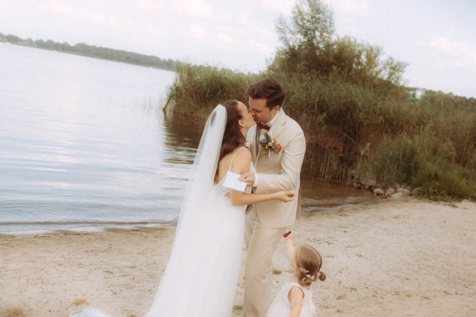 Ein Brautpaar küsst sich am Strand eines Sees, während das kleine Blumenmädchen ihre Aufmerksamkeit sucht. Ein emotionaler Moment, festgehalten von einem Hochzeitsfotograf in Schwerin und MV, perfekt für authentische Hochzeitsbilder in der Natur.