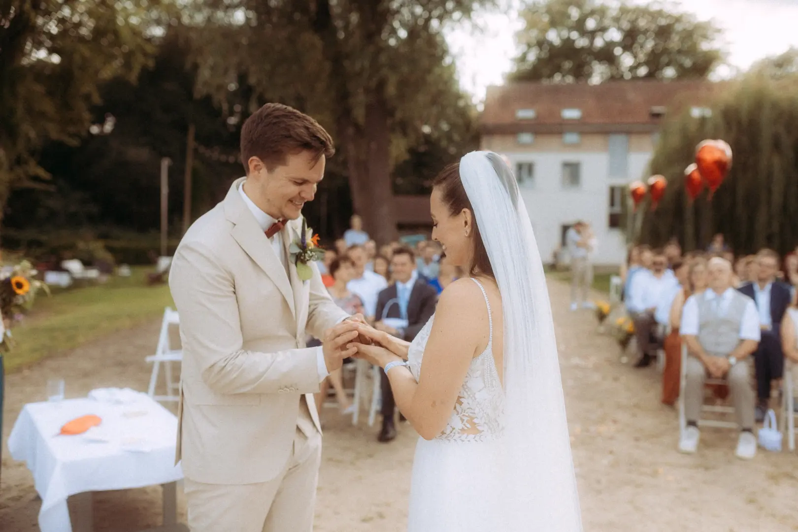 Ein Brautpaar tauscht während einer romantischen Trauung im Freien die Ringe. Der Bräutigam im beigen Anzug und die Braut im weißen Spitzenkleid mit Schleier stehen sich glücklich gegenüber, während Gäste im Hintergrund zusehen. Festliche Stimmung durch einen Hochzeitsfotograf in Schwerin und MV.